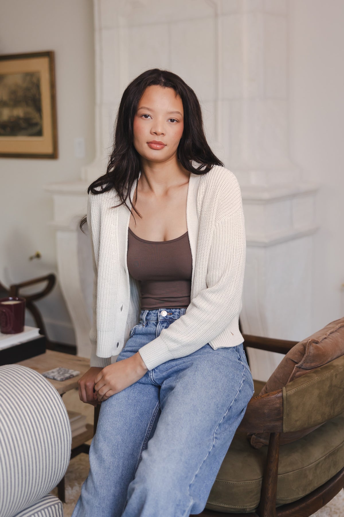 Modal sitting on sofa side wearing Chunky Knit Women's Cropped Cardigan in Oat over espresso camisole and blue jeans
