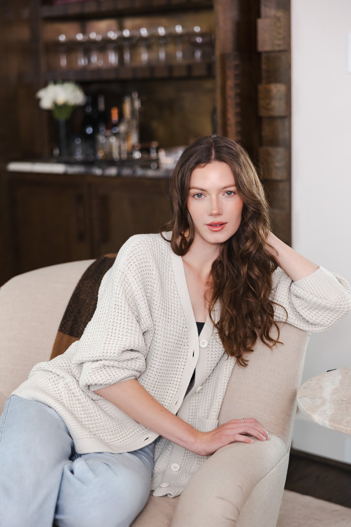 Modal wearing Chunky Knit Women's Waffle Cardigan in Oat over a blue jeans sitting on a sofa