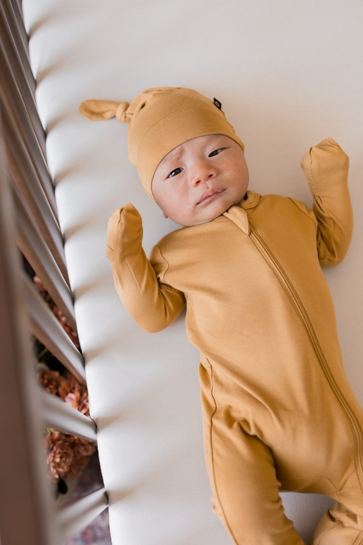 Baby wearing Knotted Cap in Ochre with zippered footie in Ochre lying in a crib