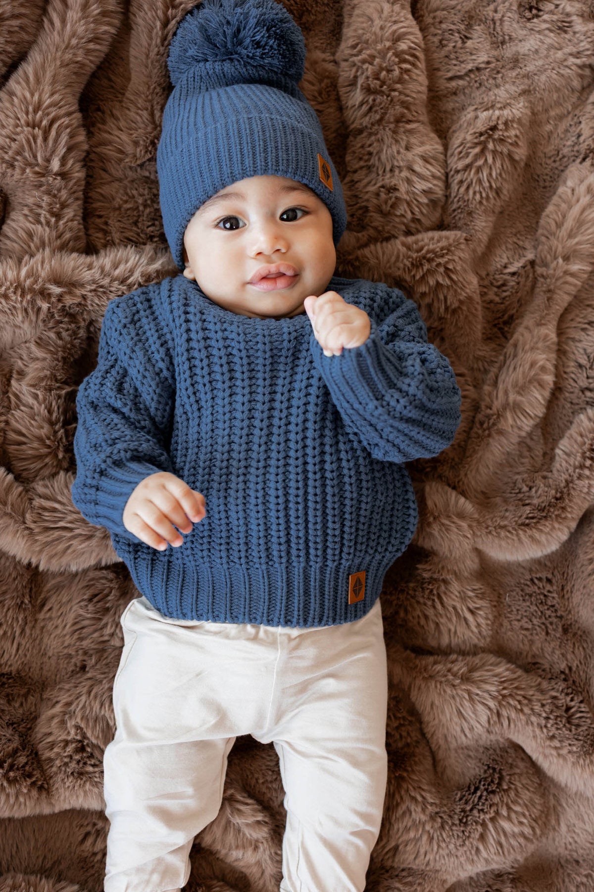 Baby wearing Chunky Knit Sweater and chunky knit pom beanie in Oxford over white pants lying on a brown blanket