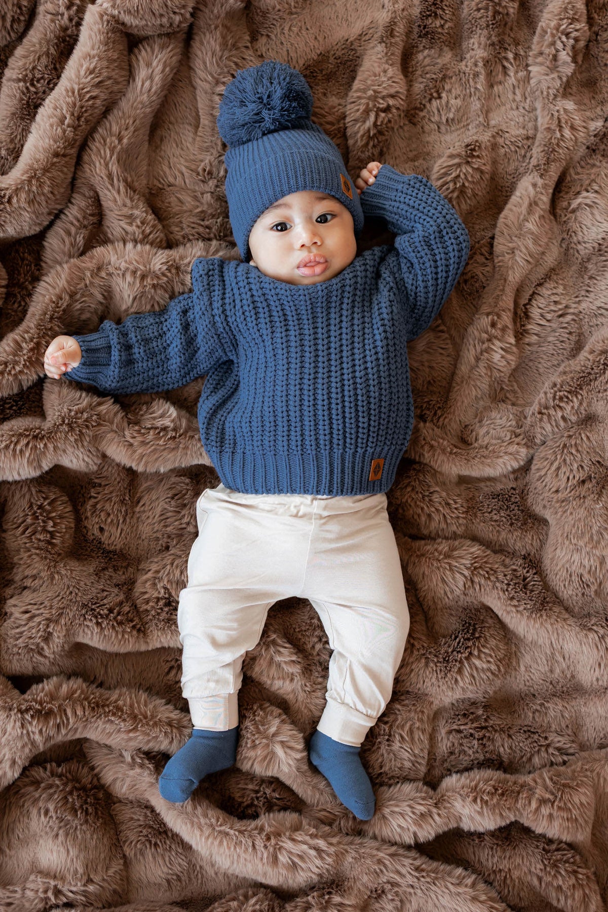 A baby lying on a brown faux fur blanket, wearing a blue ribbed knit sweater, matching blue pom-pom beanie, cream pants, and blue socks.