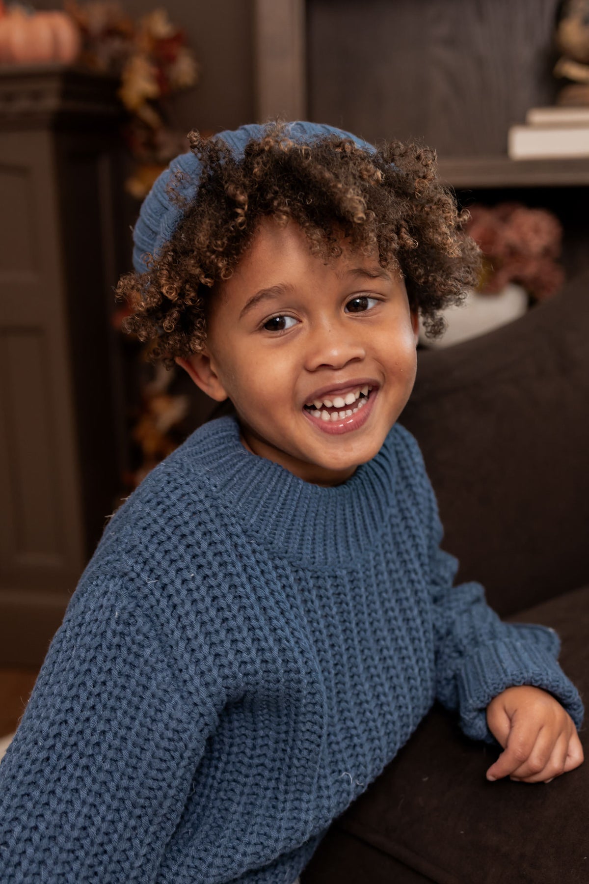 A young child with curly brown hair smiling while wearing a chunky blue knit sweater and matching blue beanie in a cozy indoor setting.