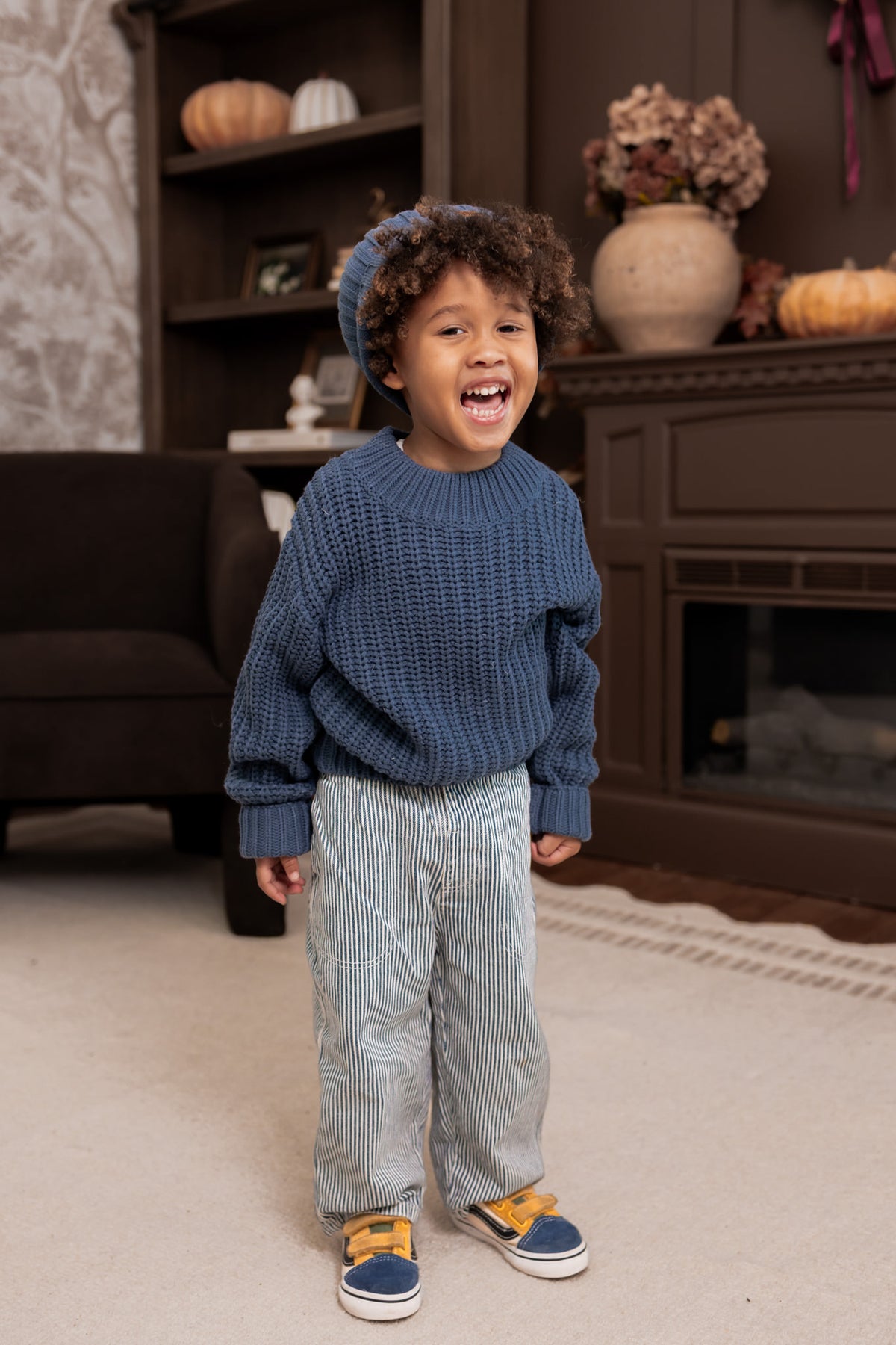 A young child wearing a chunky blue knit sweater, light blue striped pants, and blue sneakers with yellow accents, standing in a cozy living room setting.