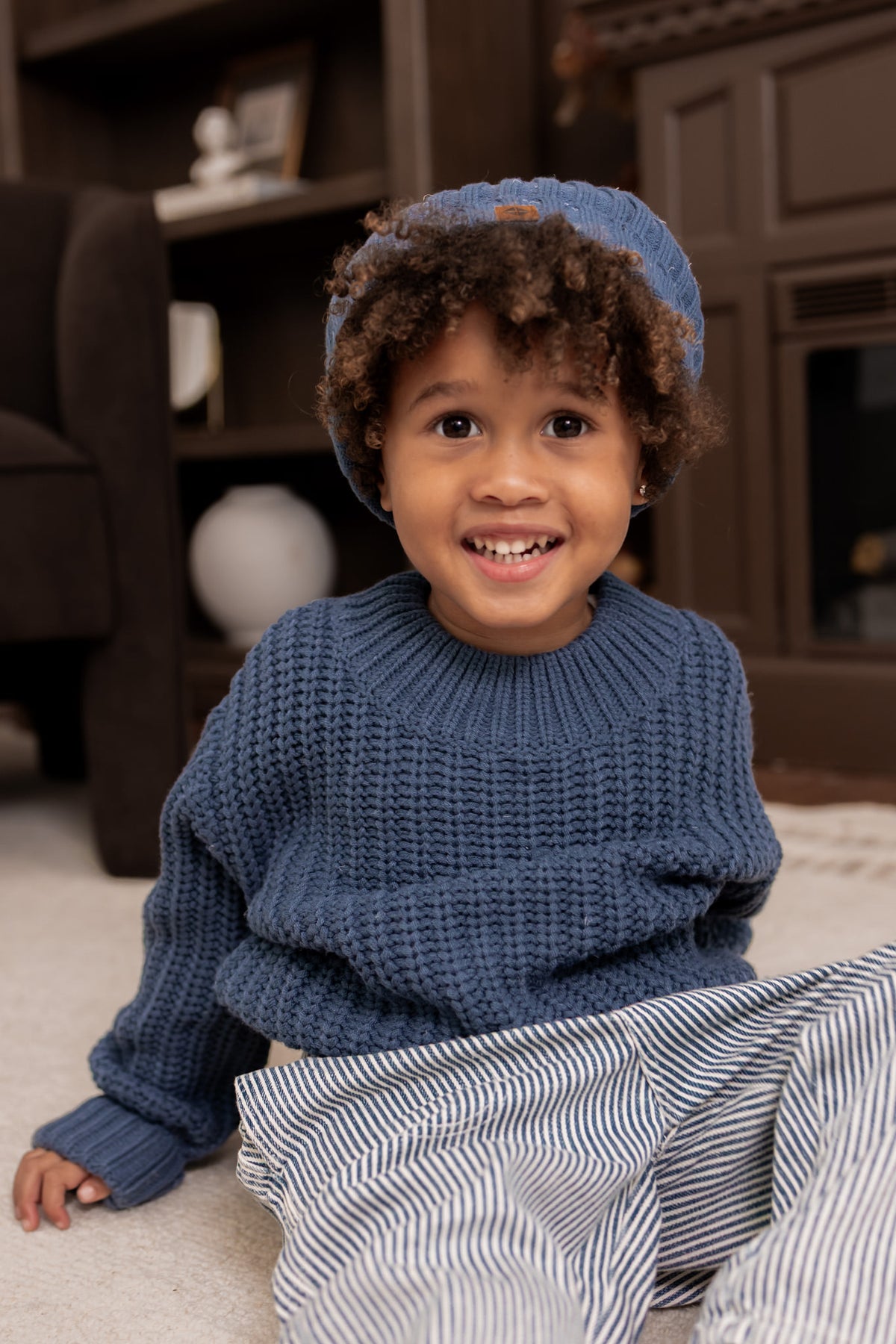 Boy wearing Chunky Knit Sweater and chunky knit pom beanie in Oxford over white pants sitting on a white carpet