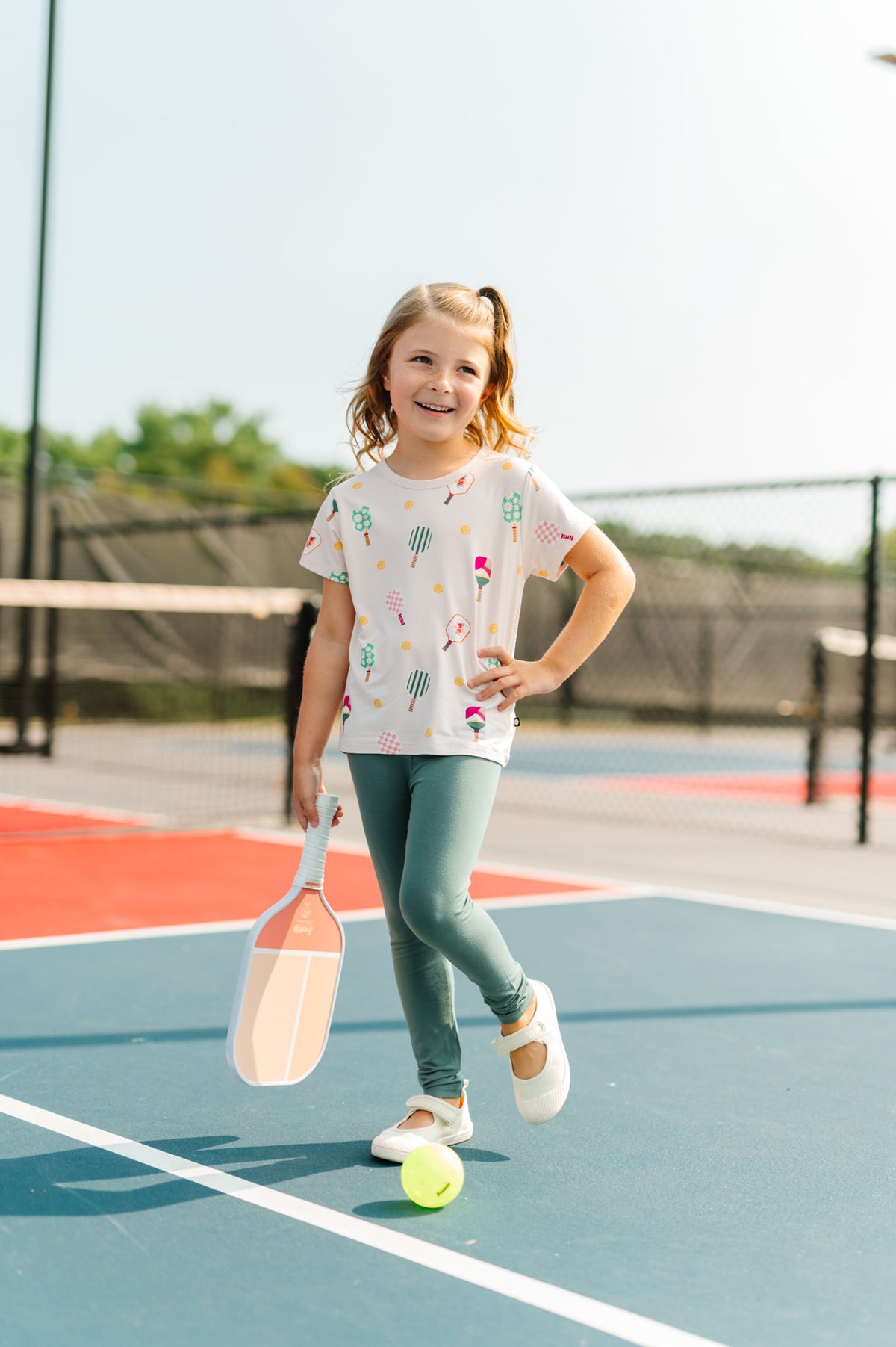 Girl posing in Toddler Basic Tee in Pickleball print over green leggings holding a bat on pickleball court