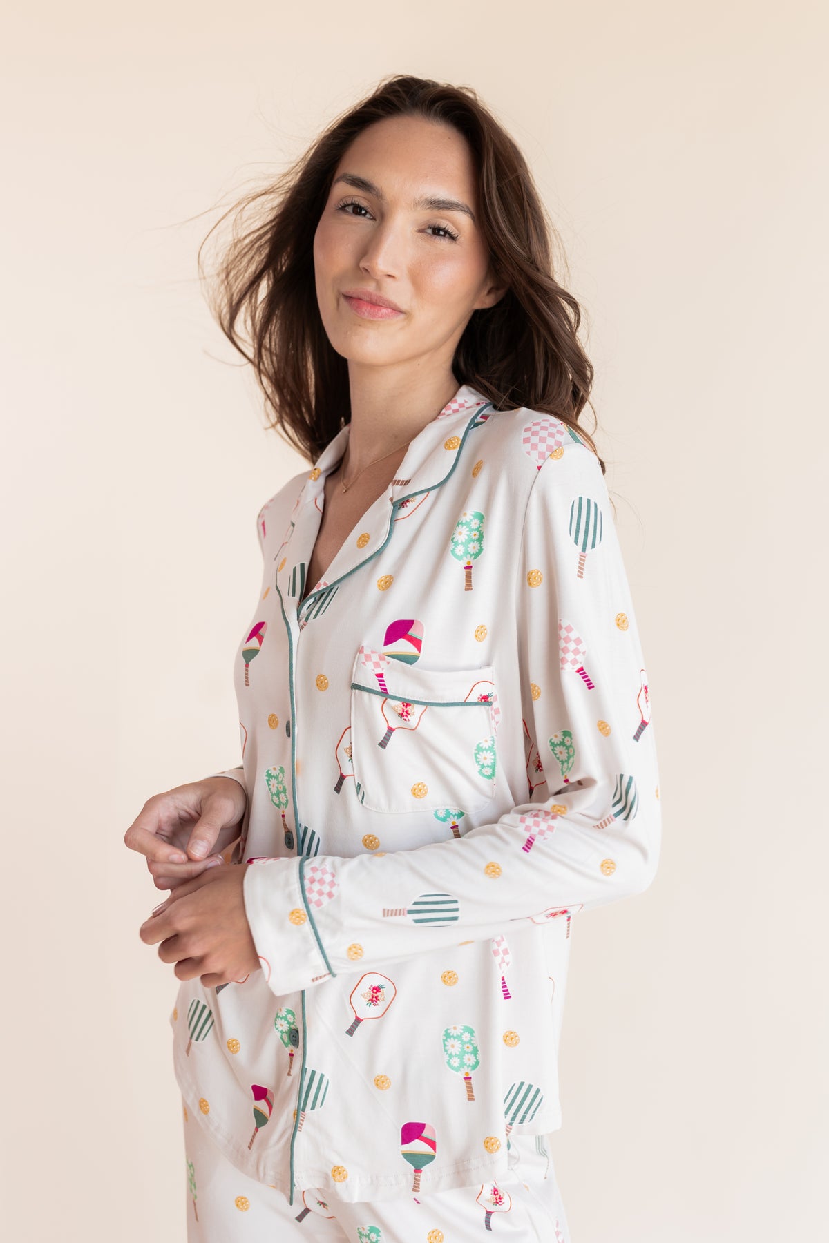 A woman wearing a long-sleeve pajama shirt with a colorful pickleball pattern on a cream background, posed against a solid light beige backdrop.