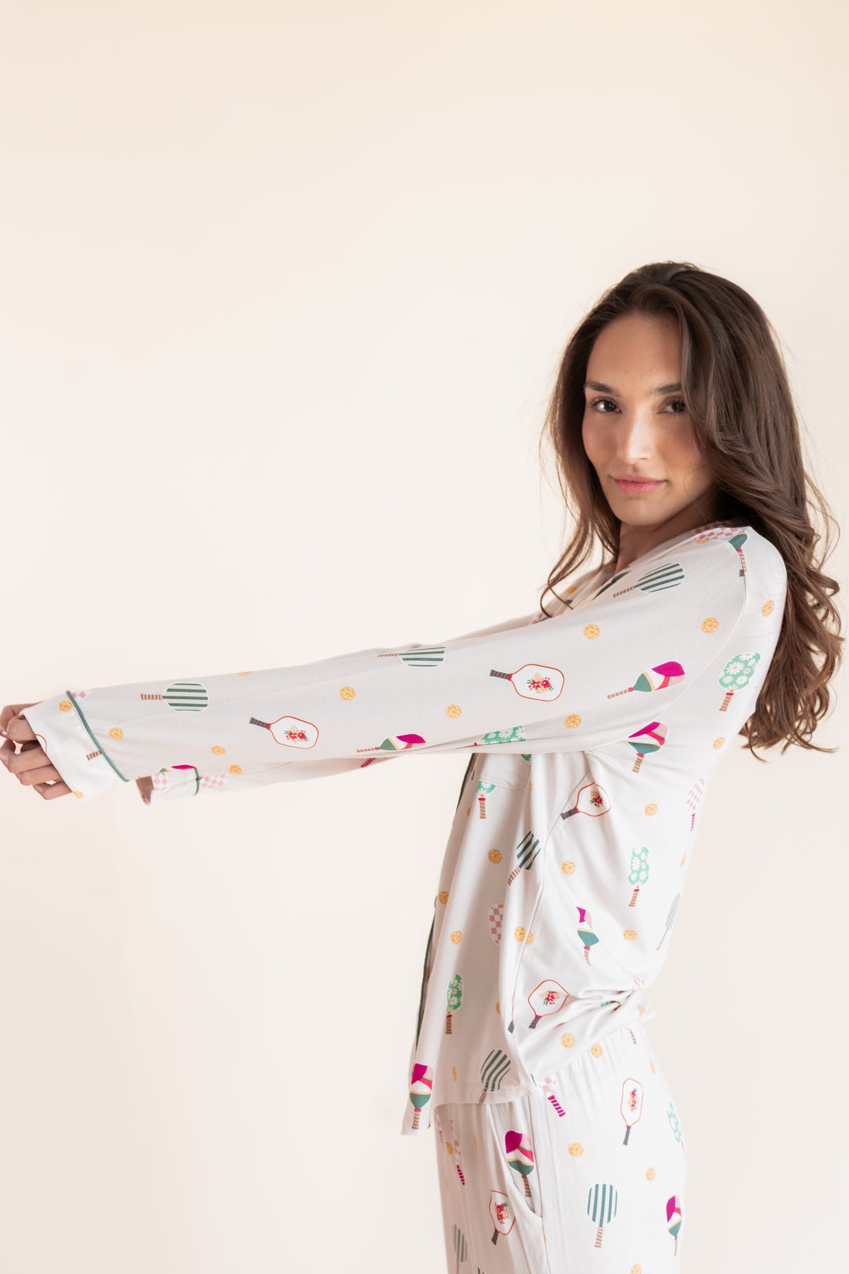 A woman modeling a long-sleeve white pajama set with a colorful pickleball print, posed against a solid light beige background.
