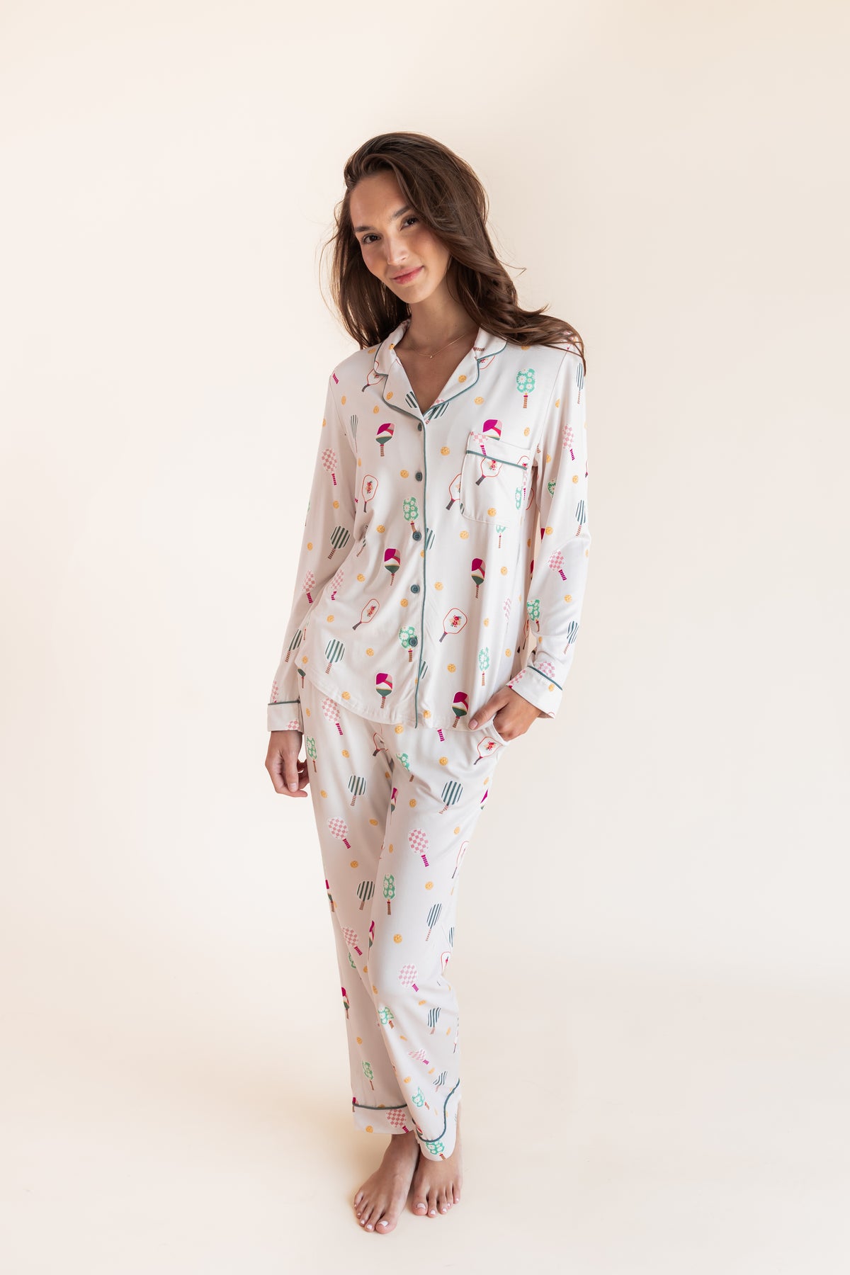 A woman modeling a matching long-sleeve pajama set with a white base and a colorful pickleball print against a plain cream studio background.