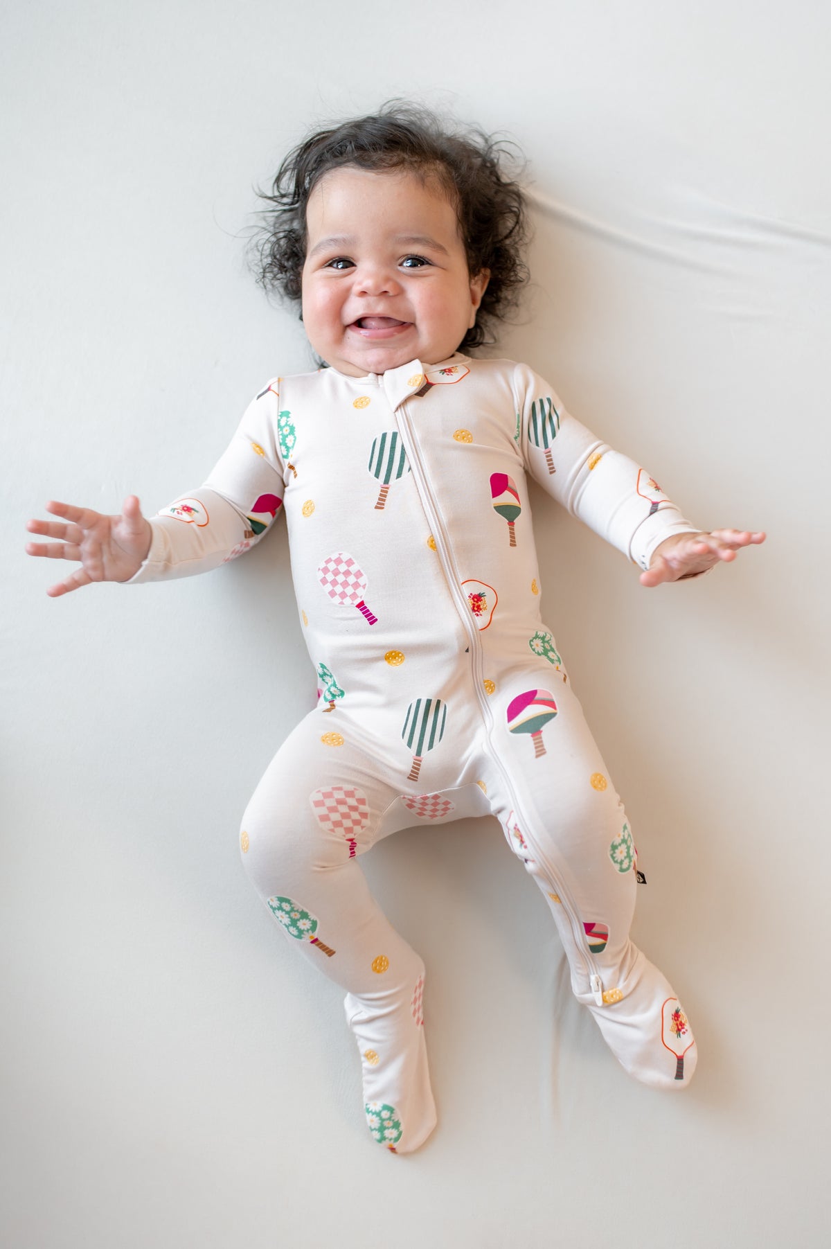 Baby wearing Zippered Footie in Pickleball print on a beige background