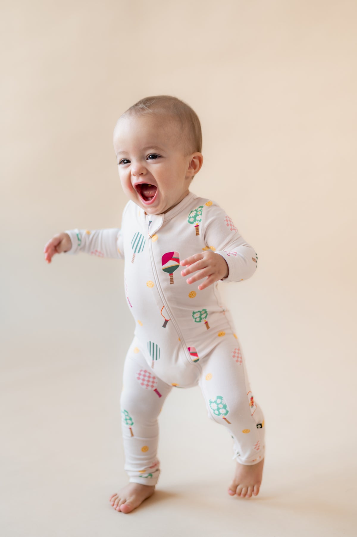 Baby wearing Zippered Romper in Pickleball print on a beige background