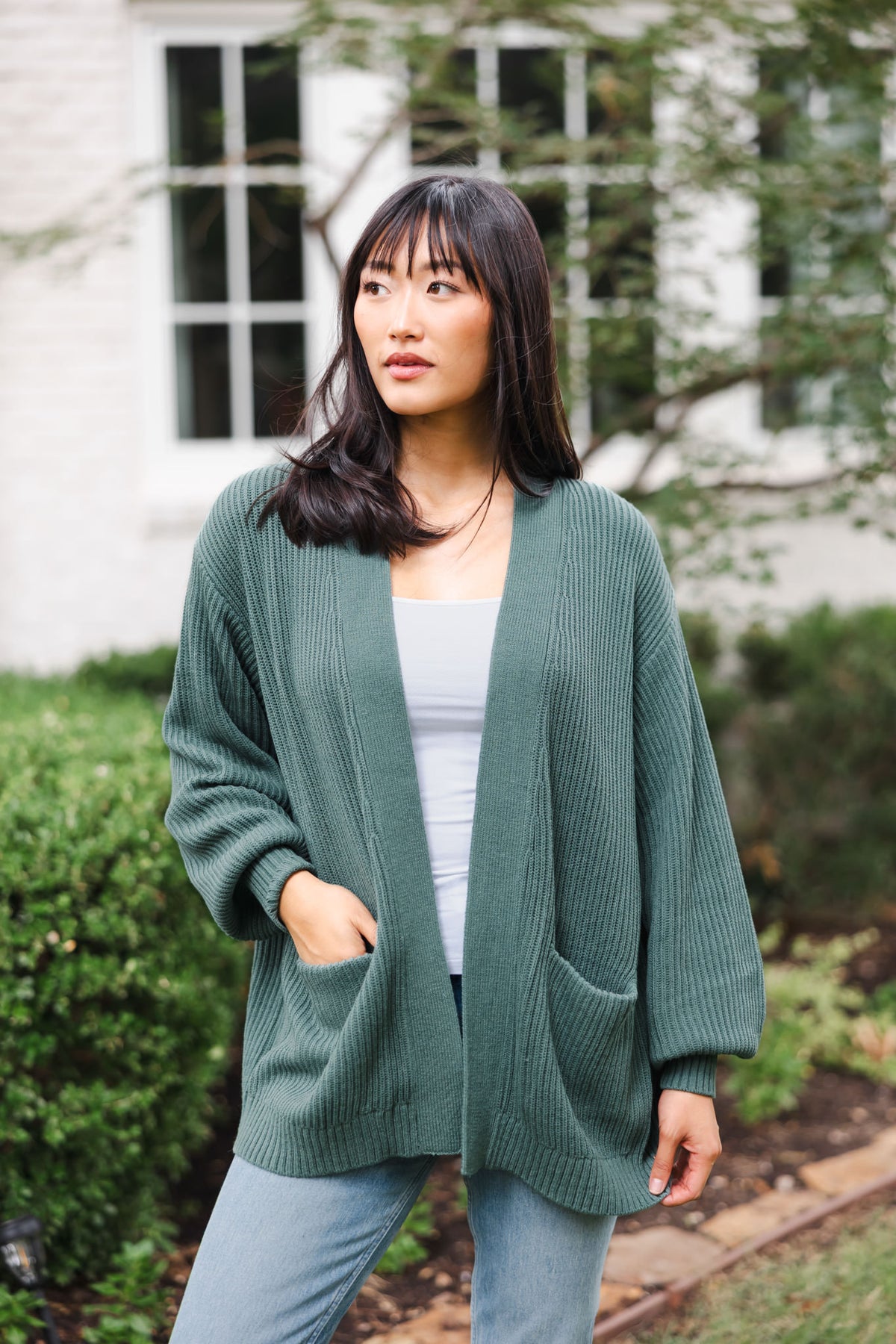Modal posing outdoors wearing Chunky knit oversized cardigan in pine over white camisole and blue jeans