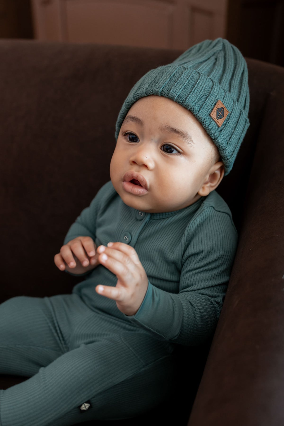 Baby wearing Chunky knit ribbed beanie in pine over pine romper on a brown sofa