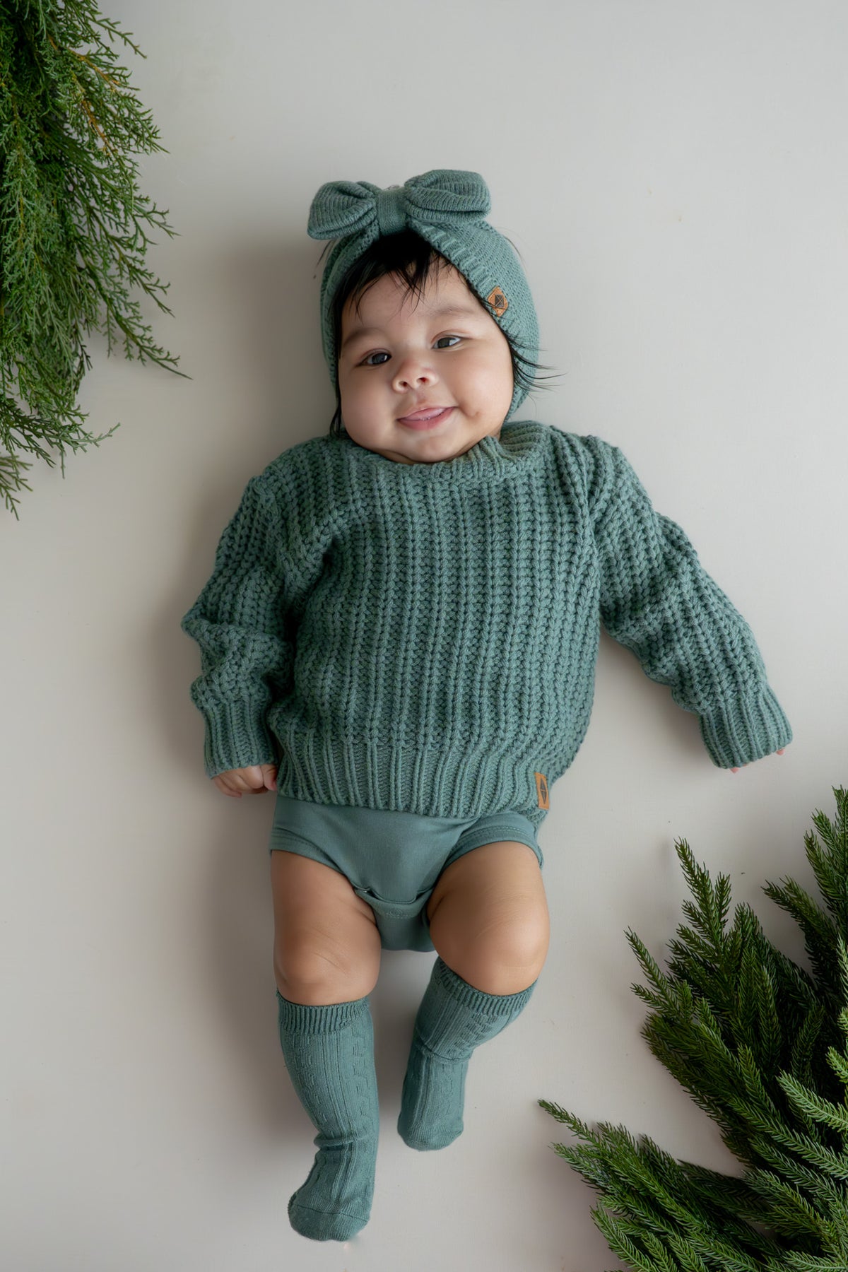 Baby wearing Chunky Knit Sweater and chunky knit bow headband in Pine lying on a beige background