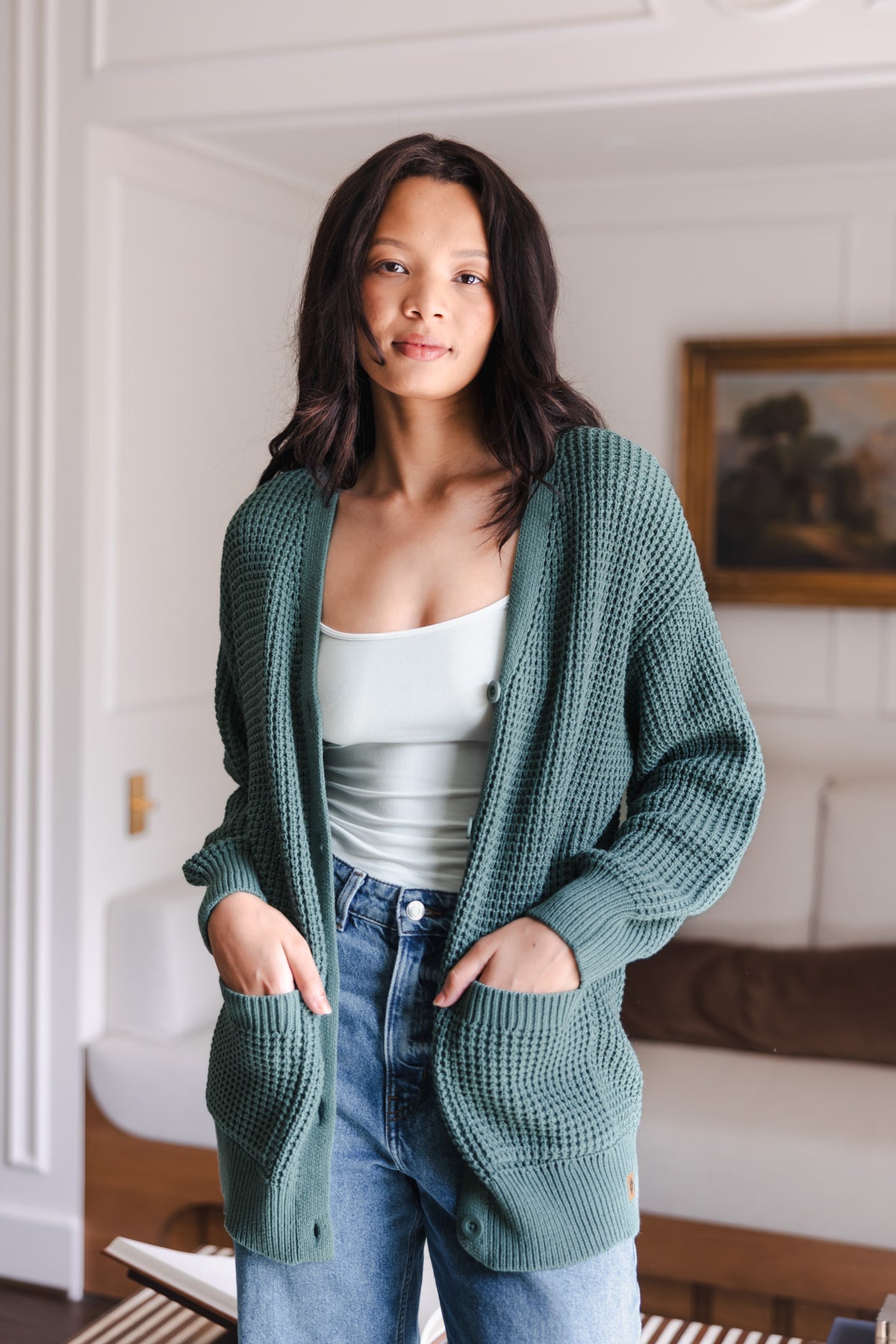 Modal wearing Chunky Knit Women's Waffle Cardigan in Pine over white camisole and blue jeans standing in a living room