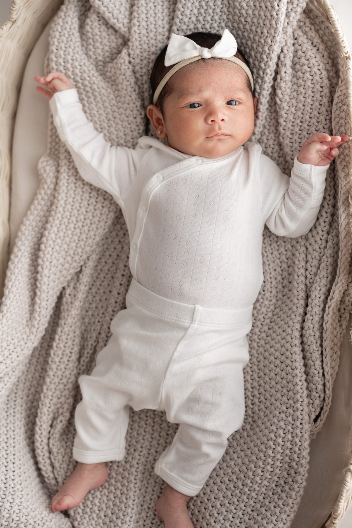 A newborn baby lying on a chunky knit beige blanket, wearing a white long-sleeve kimono and pant set and a matching white bow headband.