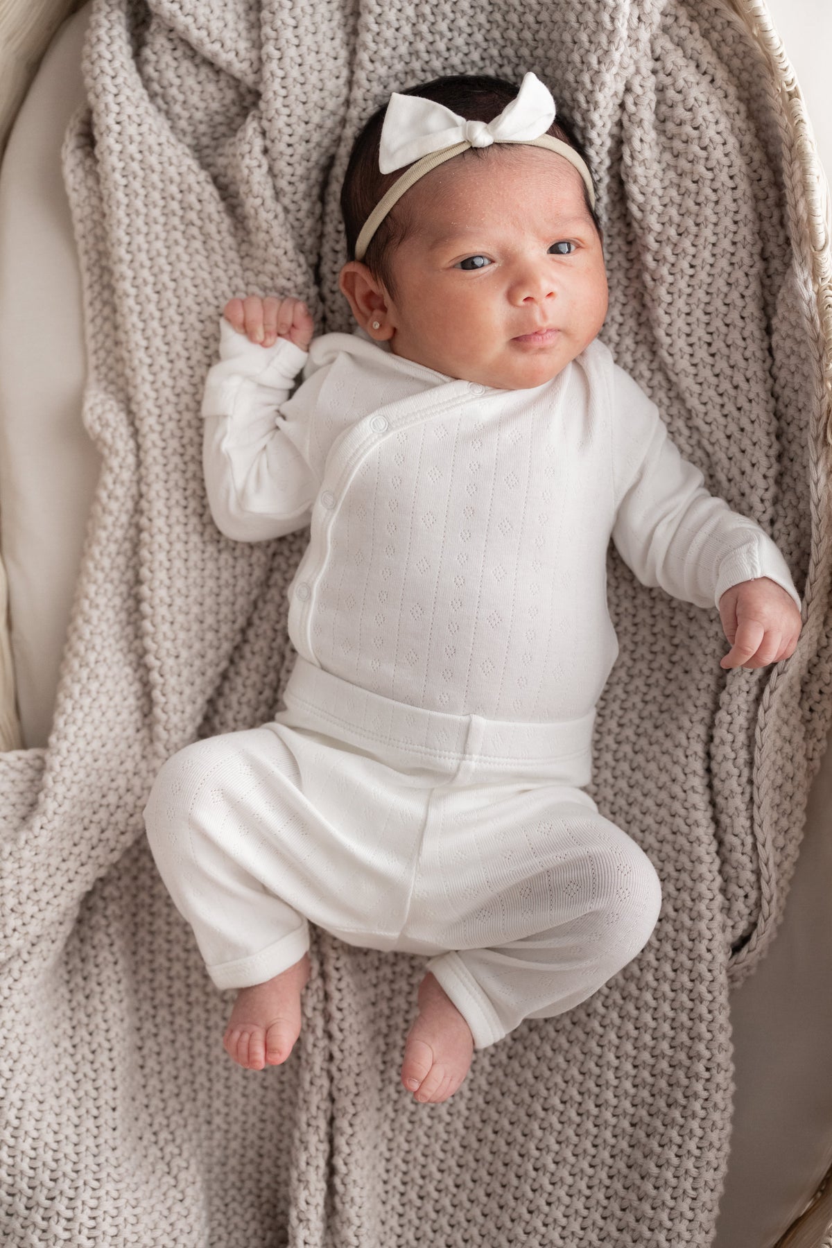 A newborn baby wearing a white long-sleeve wrap bodysuit and matching white bow headband, lying on a textured beige knit blanket.