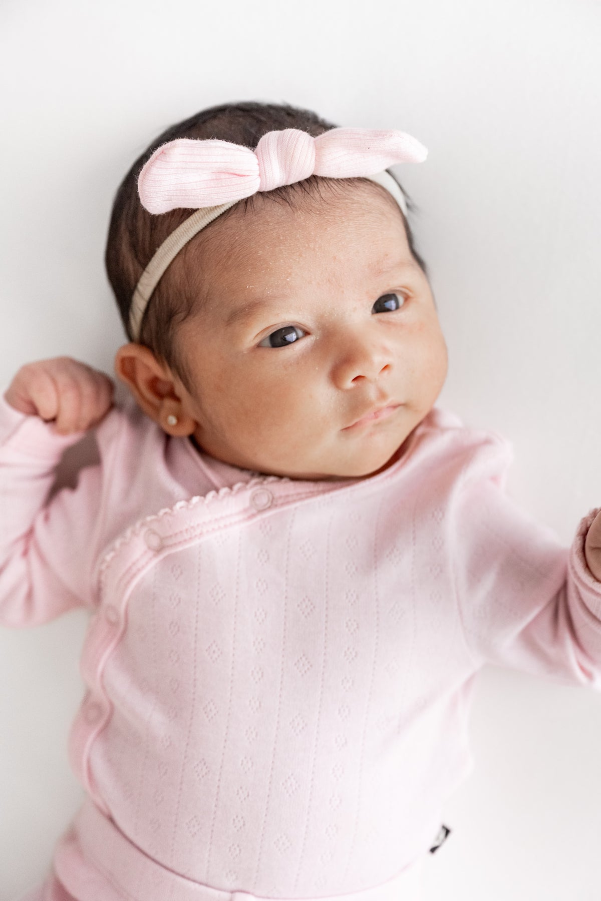 A baby wearing a light pink long-sleeve top and a matching pink bow headband, lying on a plain white background.