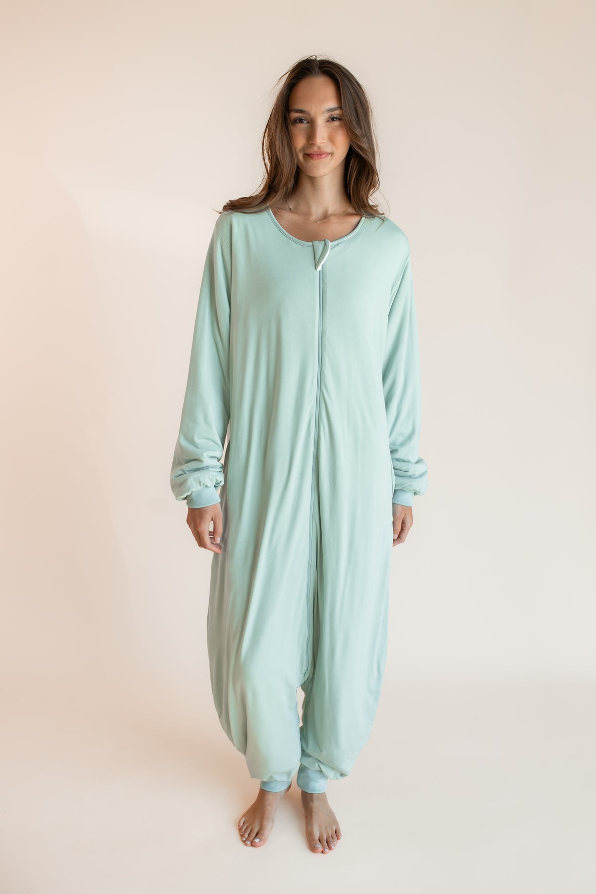 A woman modeling a light mint green one-piece sleep sack with long sleeves and a front zipper, standing against a solid pale peach background.