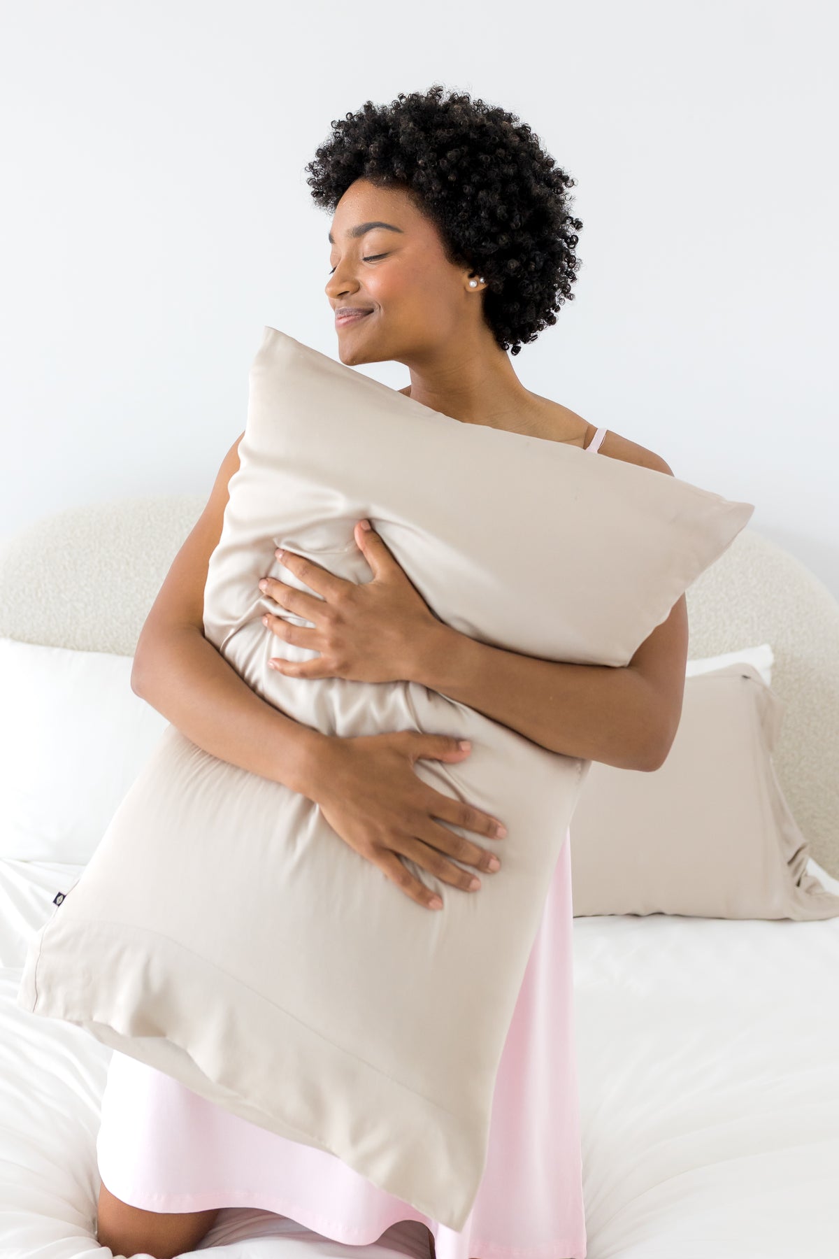 Female kneeling on a bed holding a pillow with the Bamboo Sateen Pillowcase in Sand Stone on it