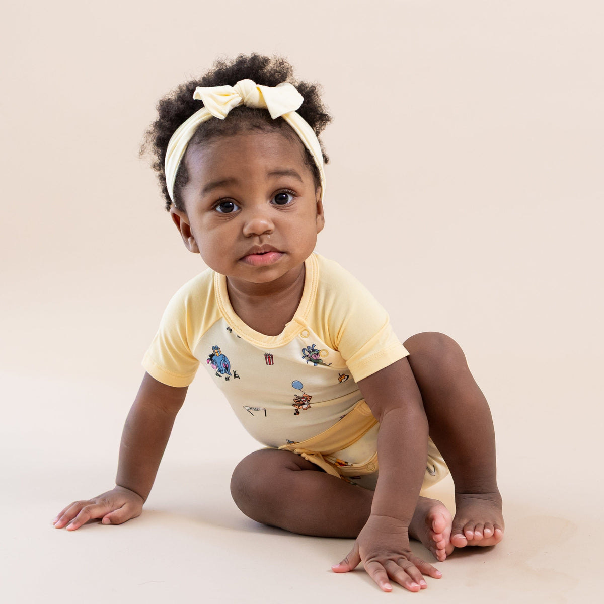 Young toddler sitting on the the floor wearing the Shortall in Winnie the Pooh Celebration with a light yellow bow