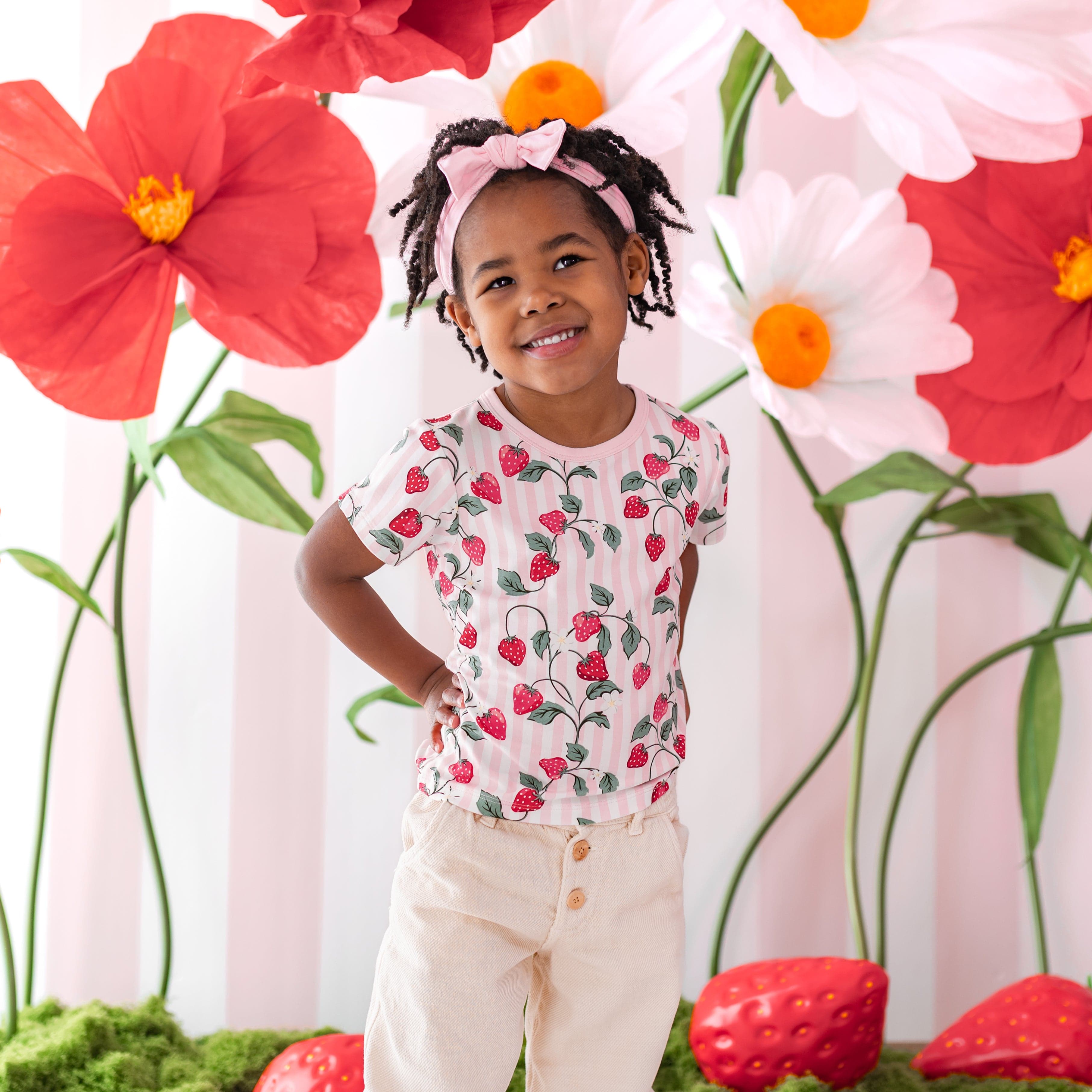 Young girl wearing the Toddler Basic Tee in Strawberry Fields standing in front of a strawberry inspired backdrop