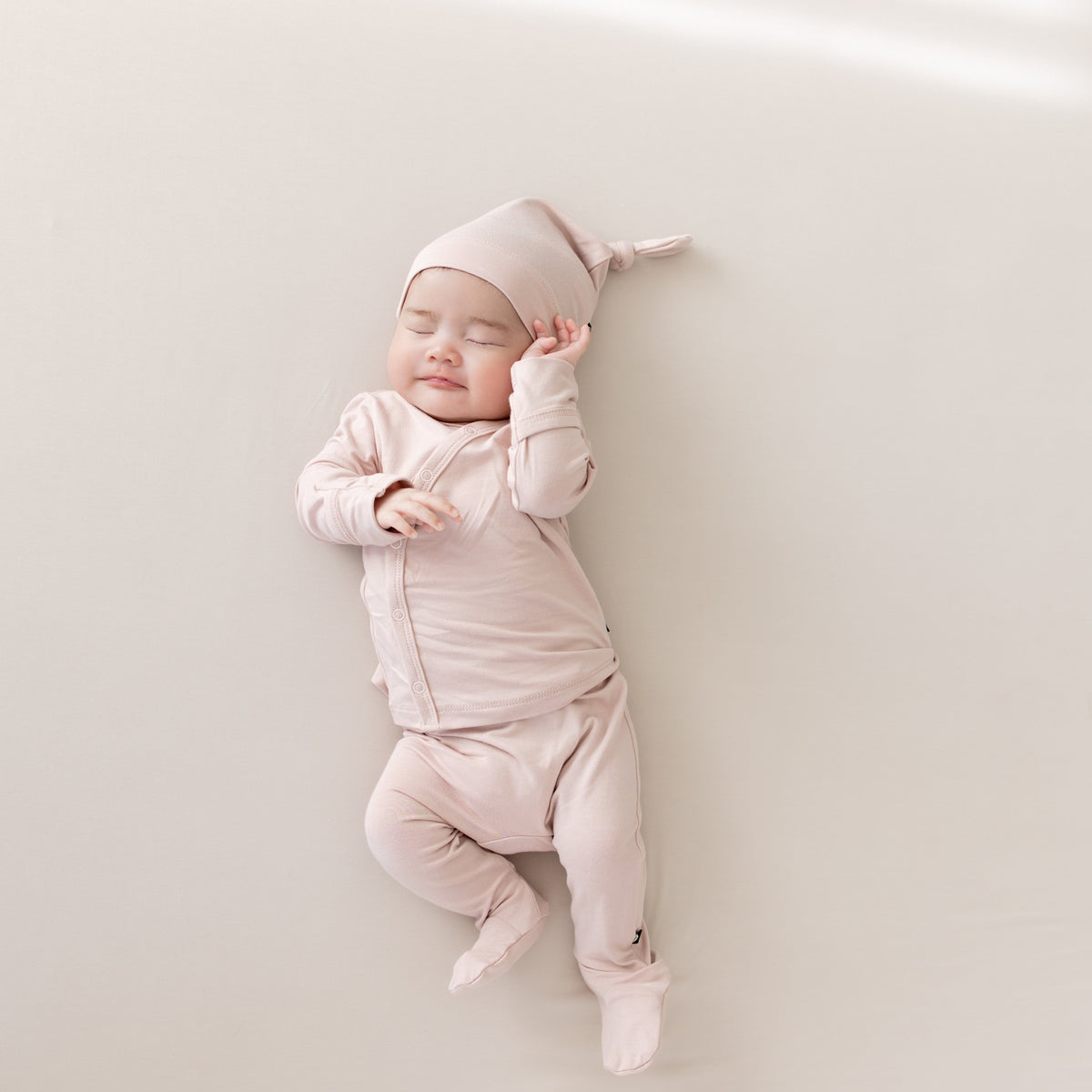Baby laying on an Oat surface while wearing the Bamboo Take Me Home Set with Cap in Blush