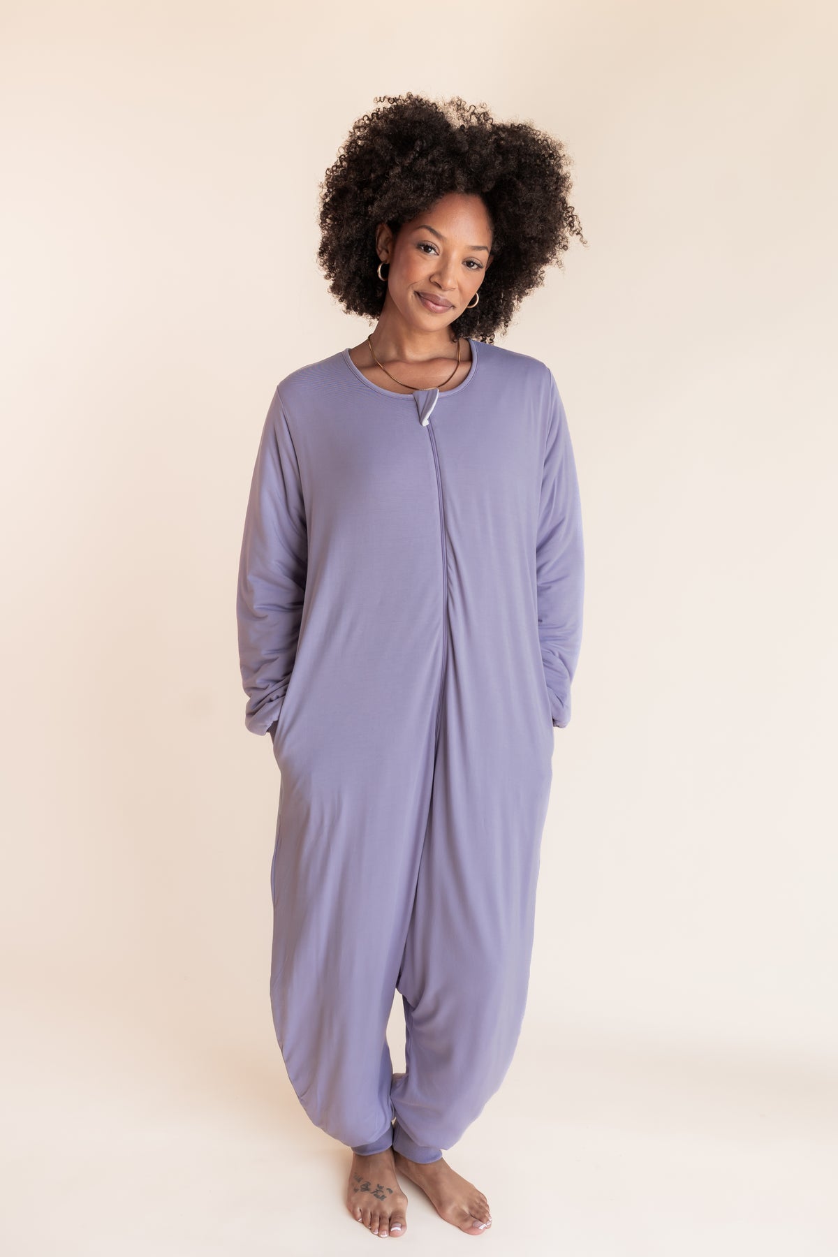 A woman with curly black hair stands barefoot against a warm cream studio background, wearing a loose, one-piece lavender sleep sack with long sleeves, side pockets, and a front zipper.