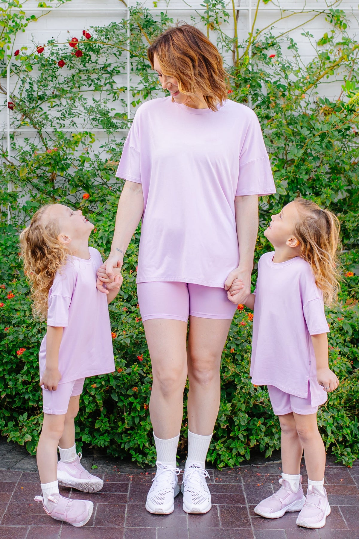 Woman and two young girls in matching lavender biker short outfits standing outdoors with greenery in the background
