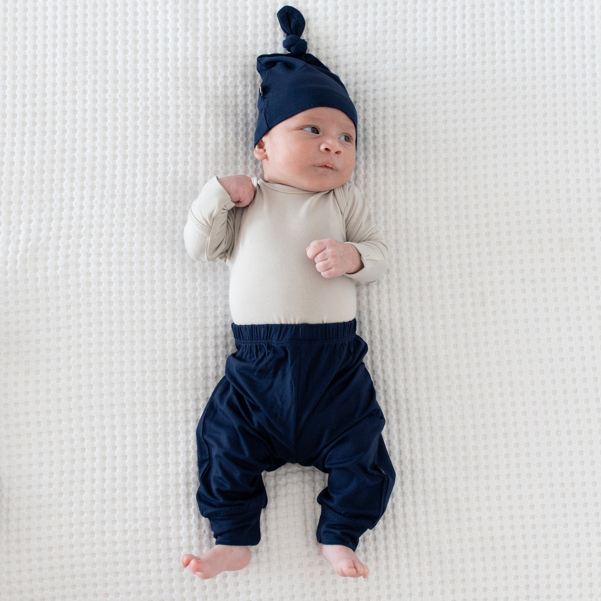 Newborn wearing Kyte Baby Knotted Cap in Navy with matching pants and a Long Sleeve Bodysuit in Oat
