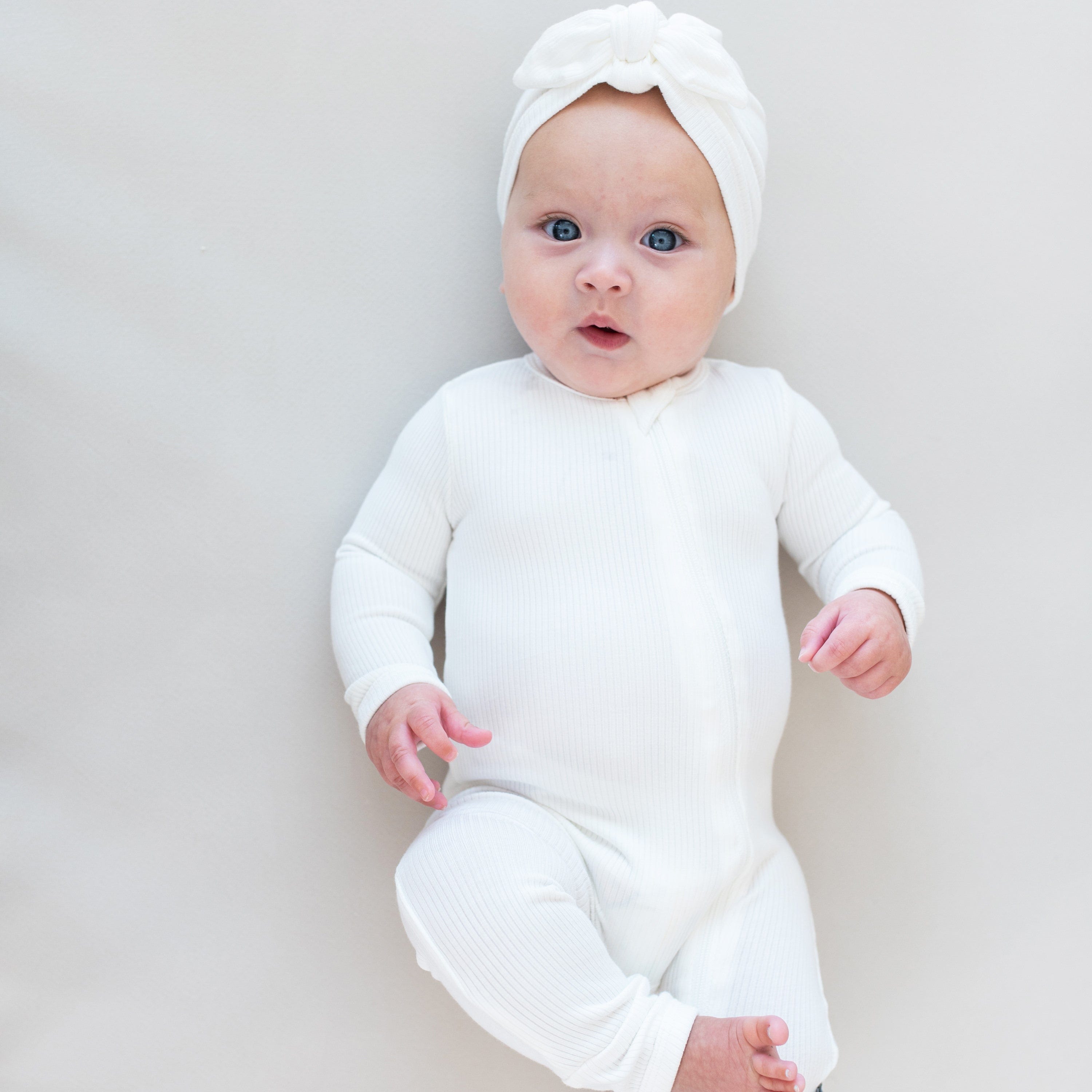 Baby laying on eggshell colored surface wearing Kyte Baby Ribbed turban style Headwrap in Cloud