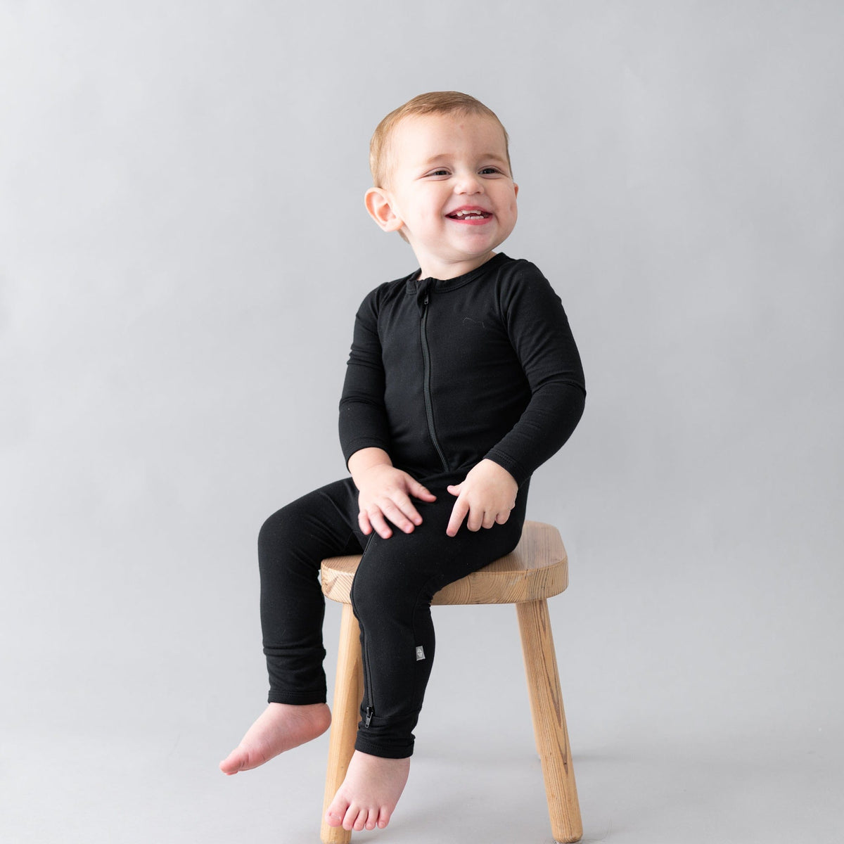 Smiling toddler sitting on a wooden stool while wearing a Zippered Romper in Midnight