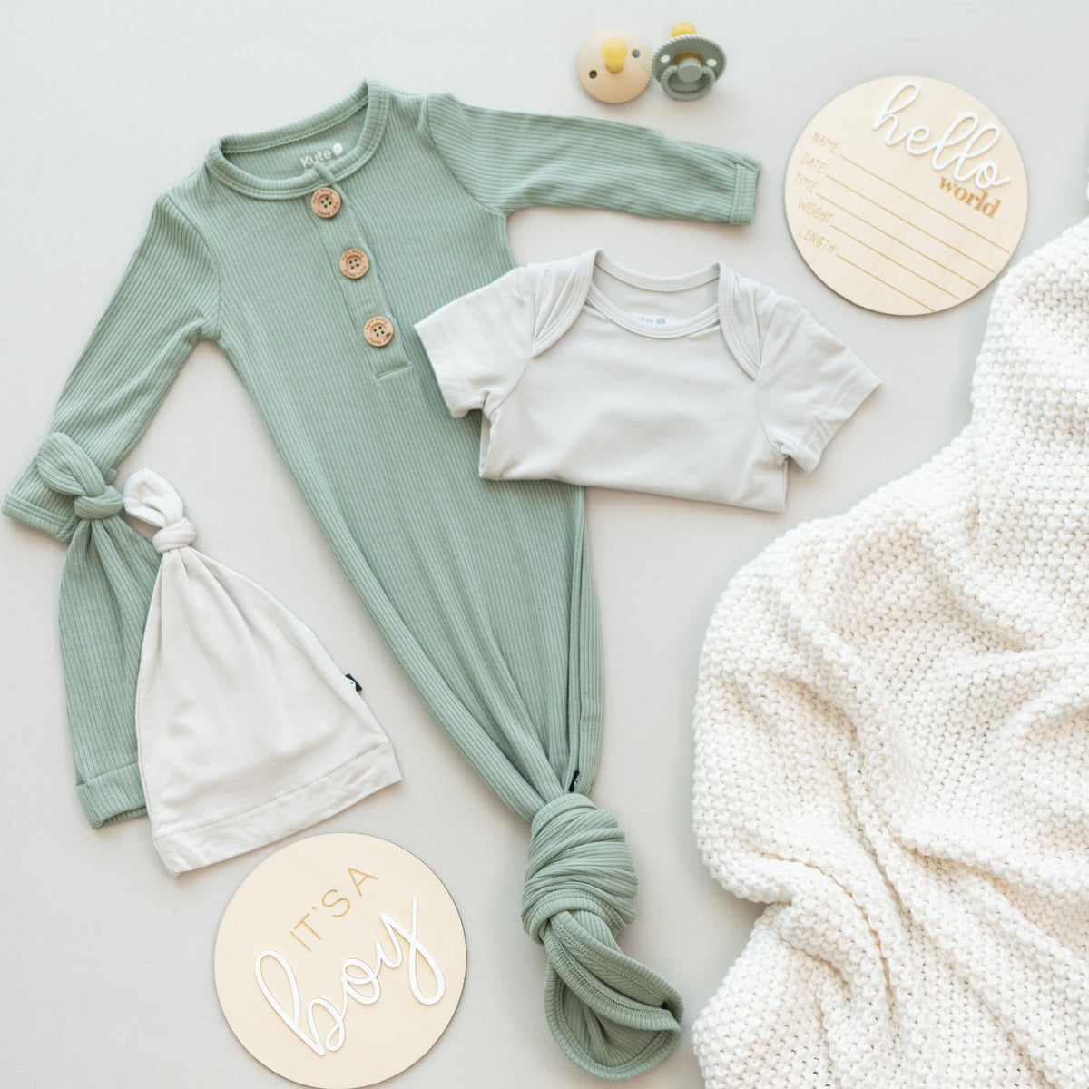 Flat Lay of Kyte Baby Products including a Knotted Gown, Short Sleeve Bodysuit, Knotted Caps, Pacis, Announcement Signs, and a Blanket
