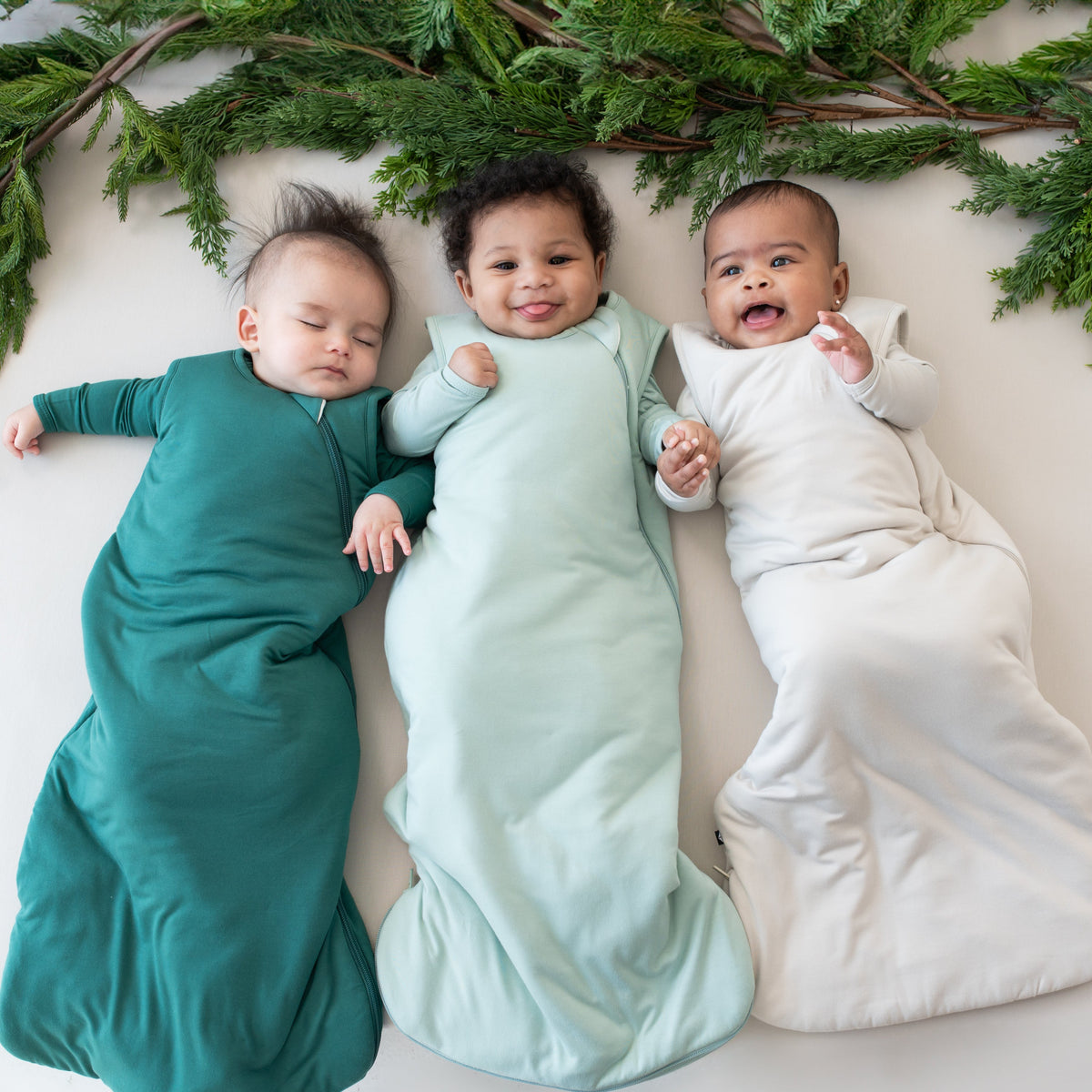 Three babies laying down wearing sleep bags in colors storm, sage and slate