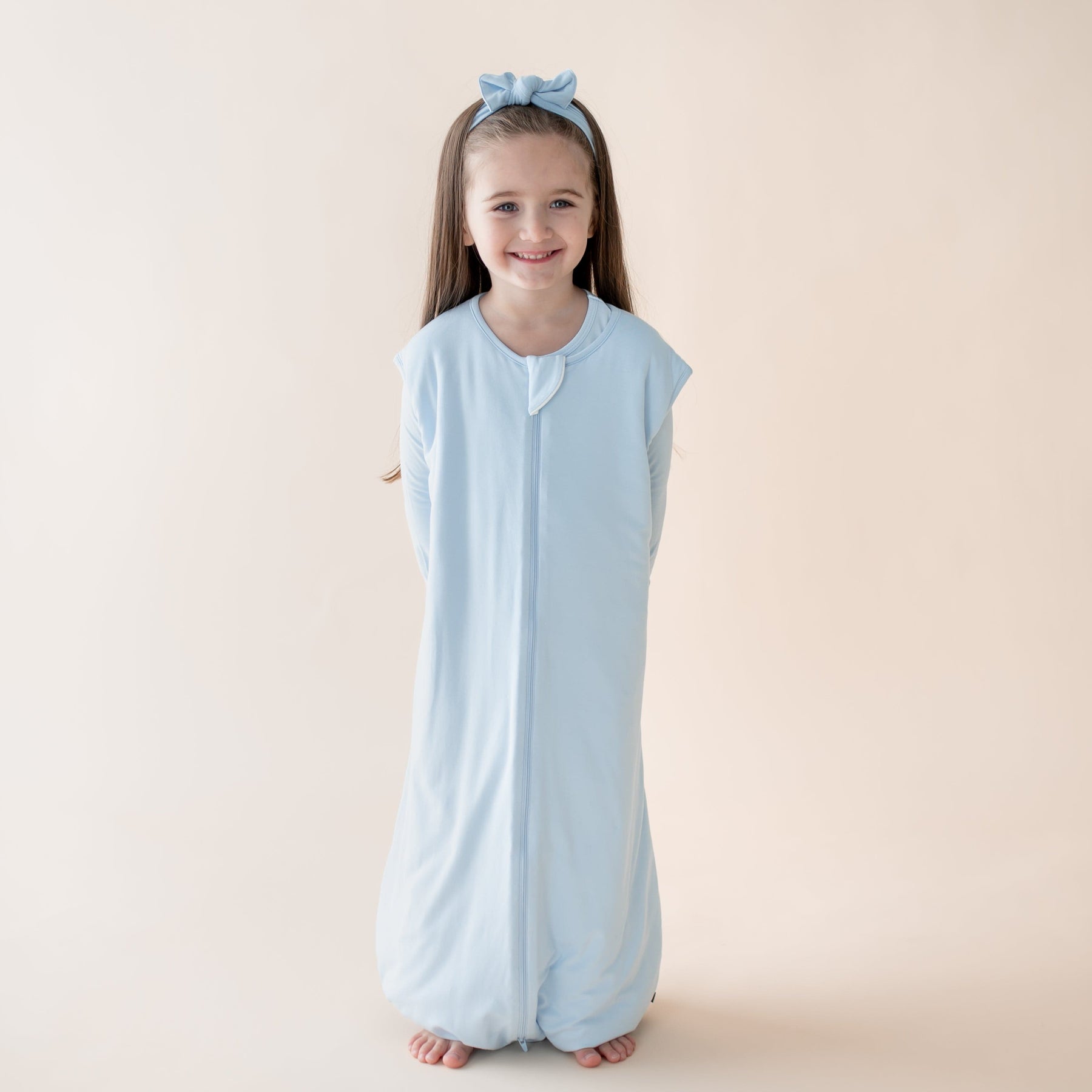 Young girl wearing a light blue sleeveless sleep bag walker with a zipper on a beige background