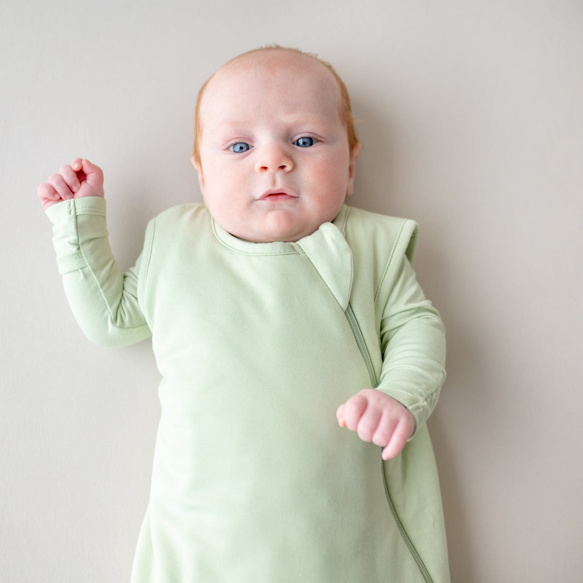 Baby wearing a light green sleep bag and footie against a plain background