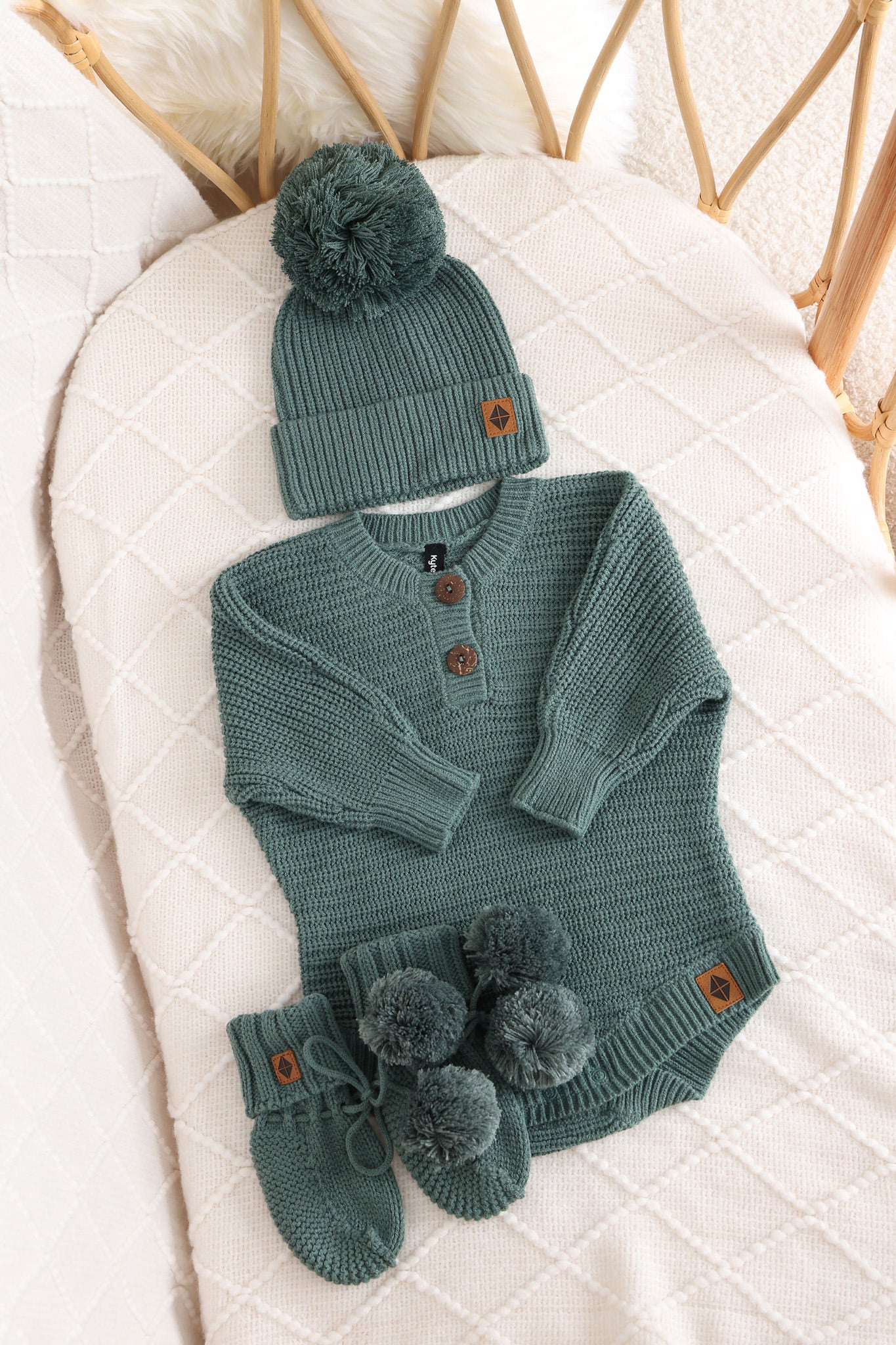 Chunky knit pom beanie, chunky knit romper and knit booties in pine laid in a crib
