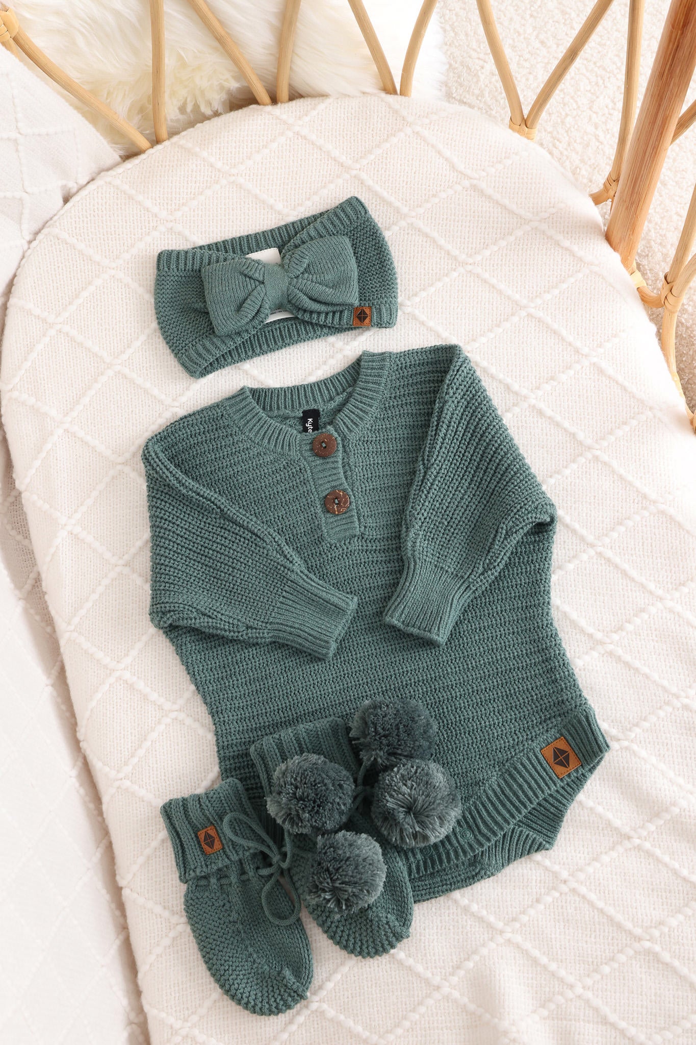 Chunky knit bow headband, chunky knit romper and knit booties in pine laid in a crib