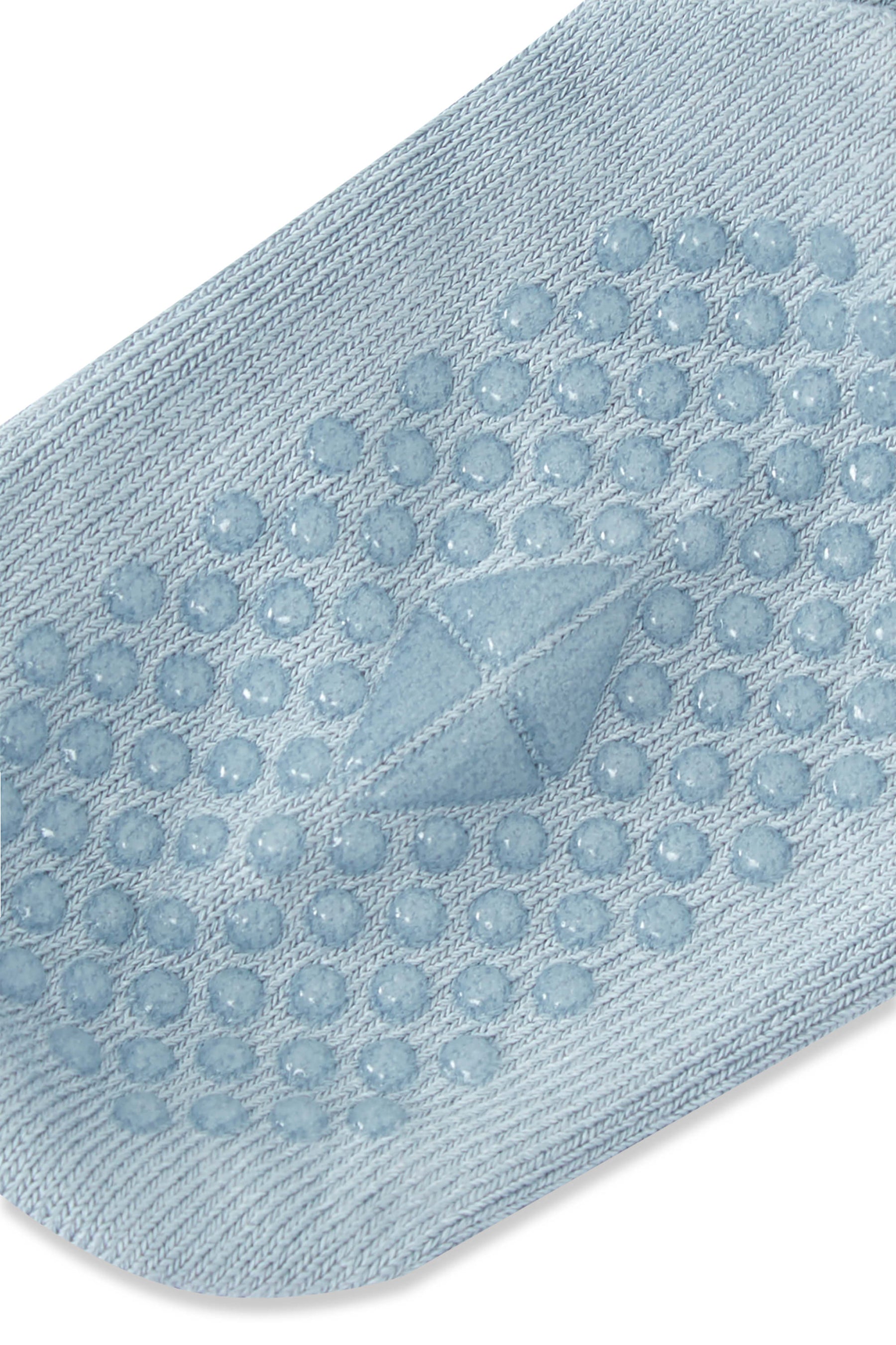 A close-up of a light blue knit sock with raised circular grip dots and a central triangular textured detail on the sole, shown on a white background.