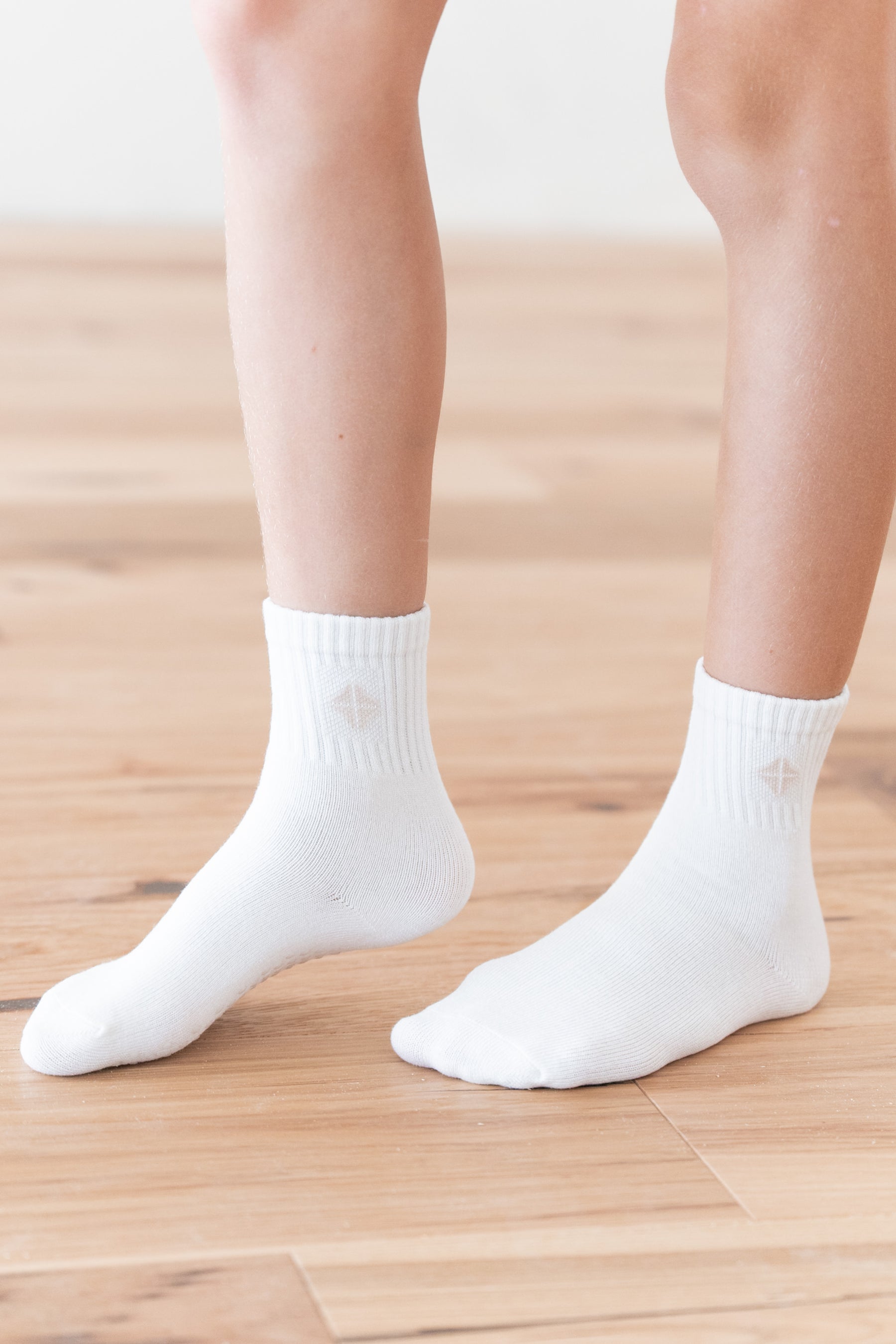 A close-up of a person's feet and lower legs wearing plain white ankle socks on a light wood floor.