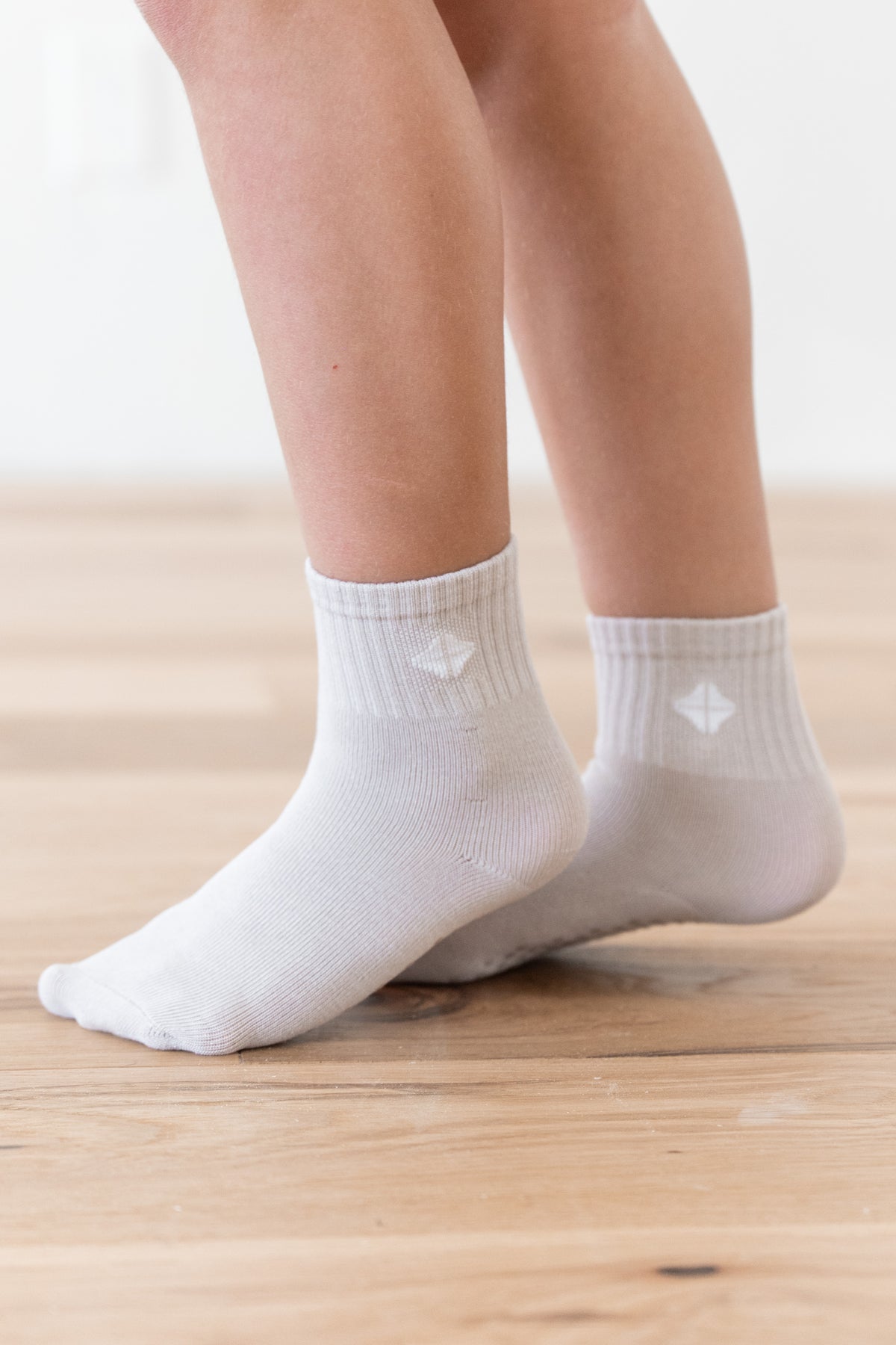 White ankle socks worn on a child's feet, standing on a light wooden floor against a bright white background.