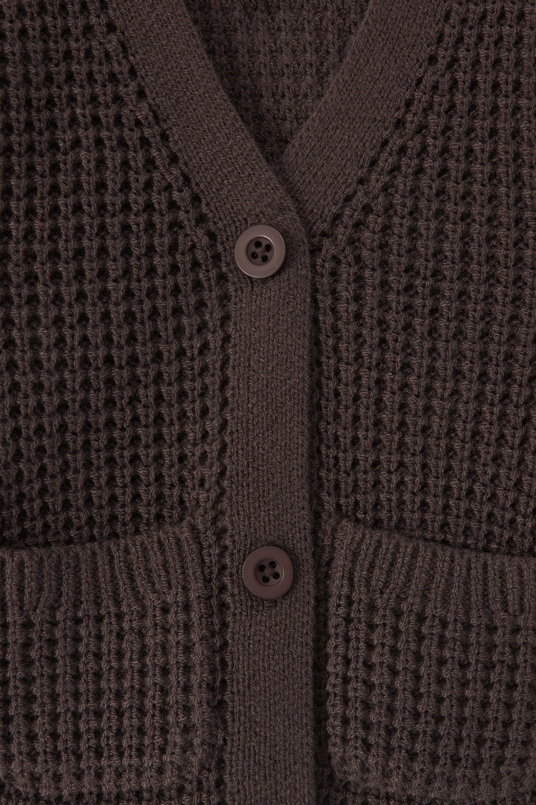 Close-up of a textured knit cardigan in a dark brown color, featuring a waffle-weave pattern, ribbed trim, and two white buttons.