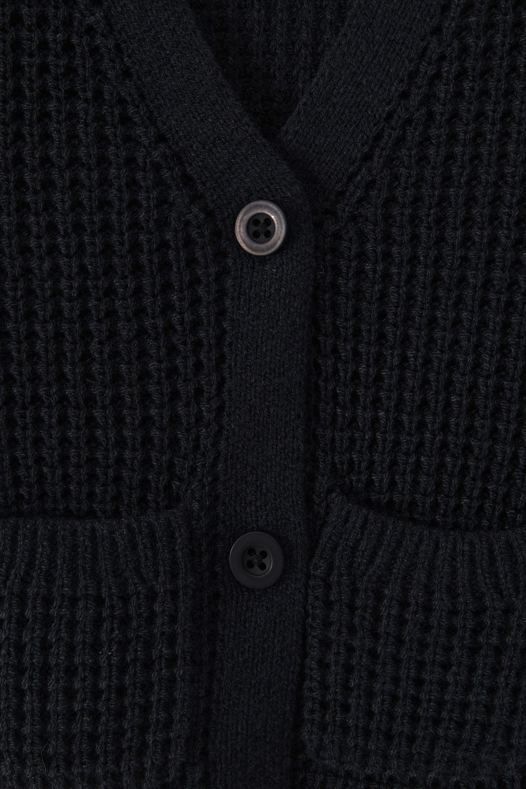 Close-up of a textured knit cardigan in black color, featuring a waffle-weave pattern, ribbed trim, and two white buttons.