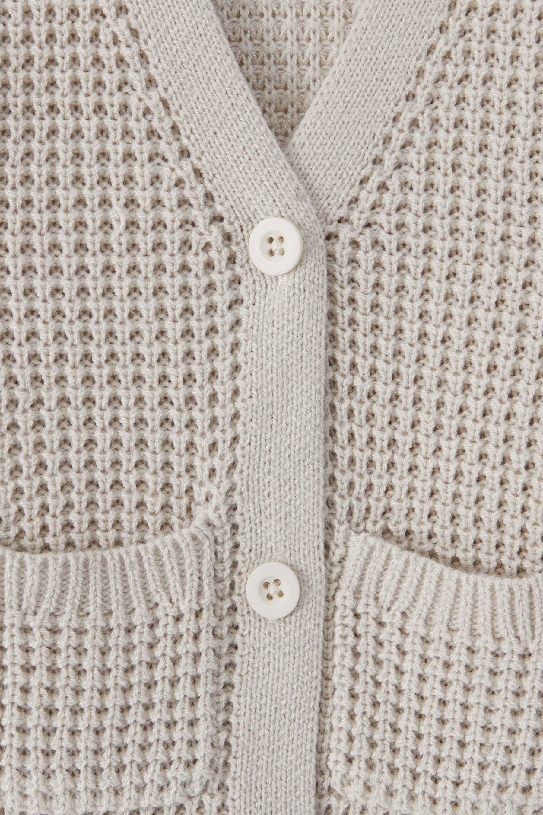 Close-up of a textured knit cardigan in an oat color, featuring a waffle-weave pattern, ribbed trim, and two white buttons.