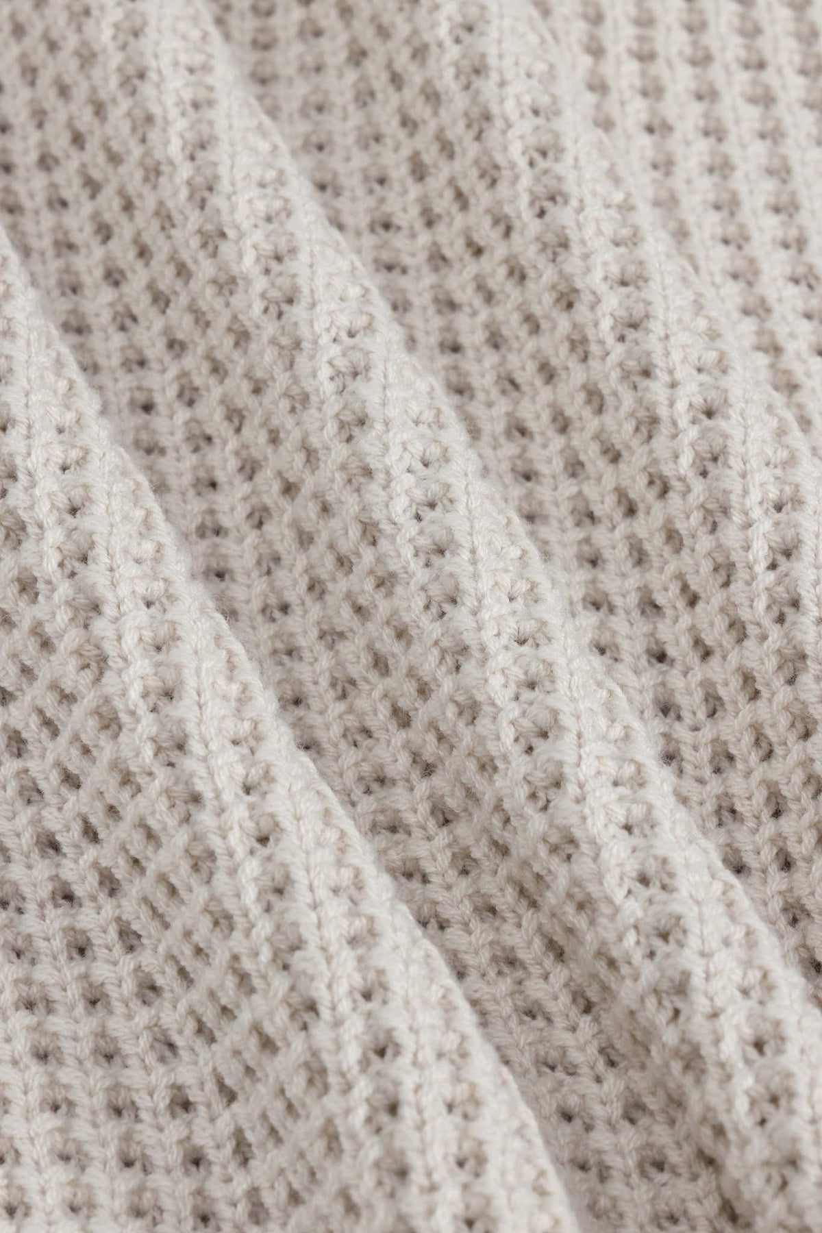 Close-up of a textured knit fabric in a warm off-white or cream tone, showing the detailed weave and soft folds.
