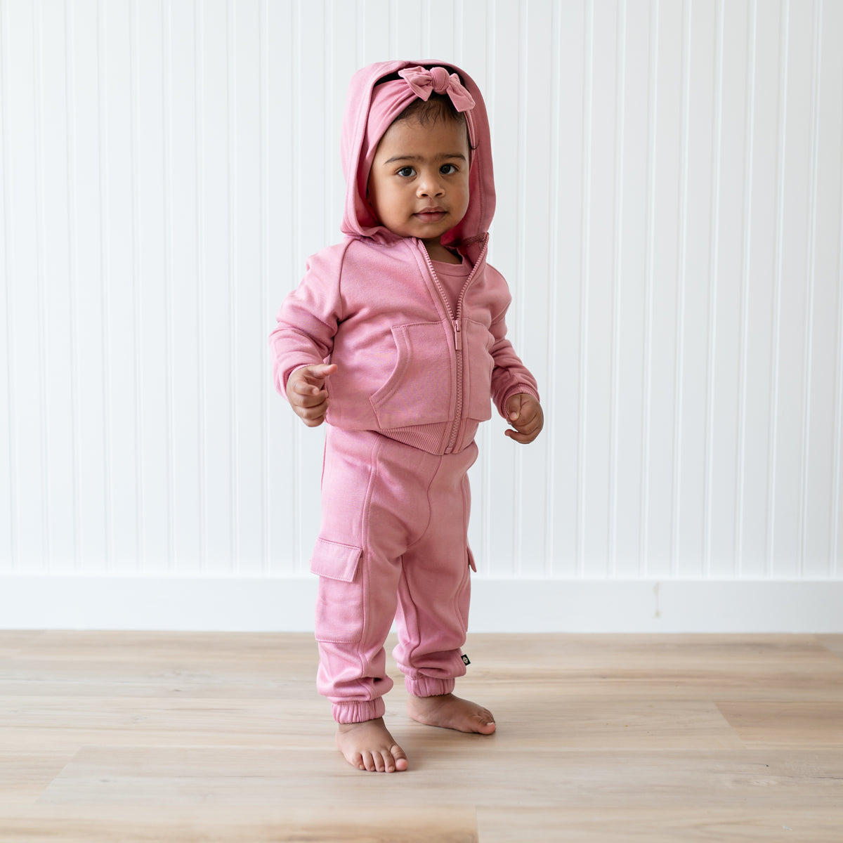 Young toddler wearing the Fleece Cargo Pants in Apple Blossom with matching fleece zip hoodie standing in front of a white paneled wall