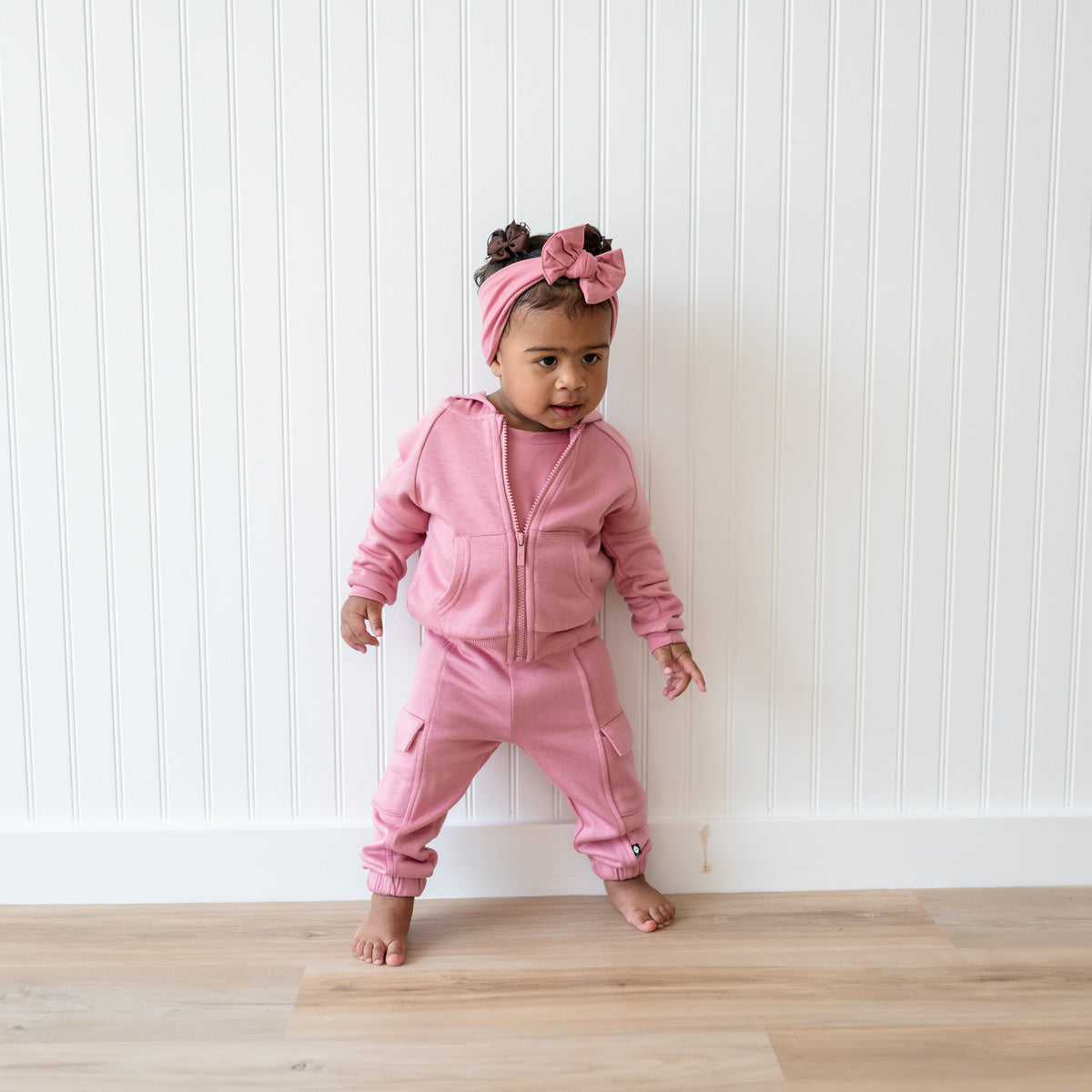 Young toddler standing in front of a white paneled wall wearing the Fleece Cargo Pants in Apple Blossom with matching fleece zip hoodie
