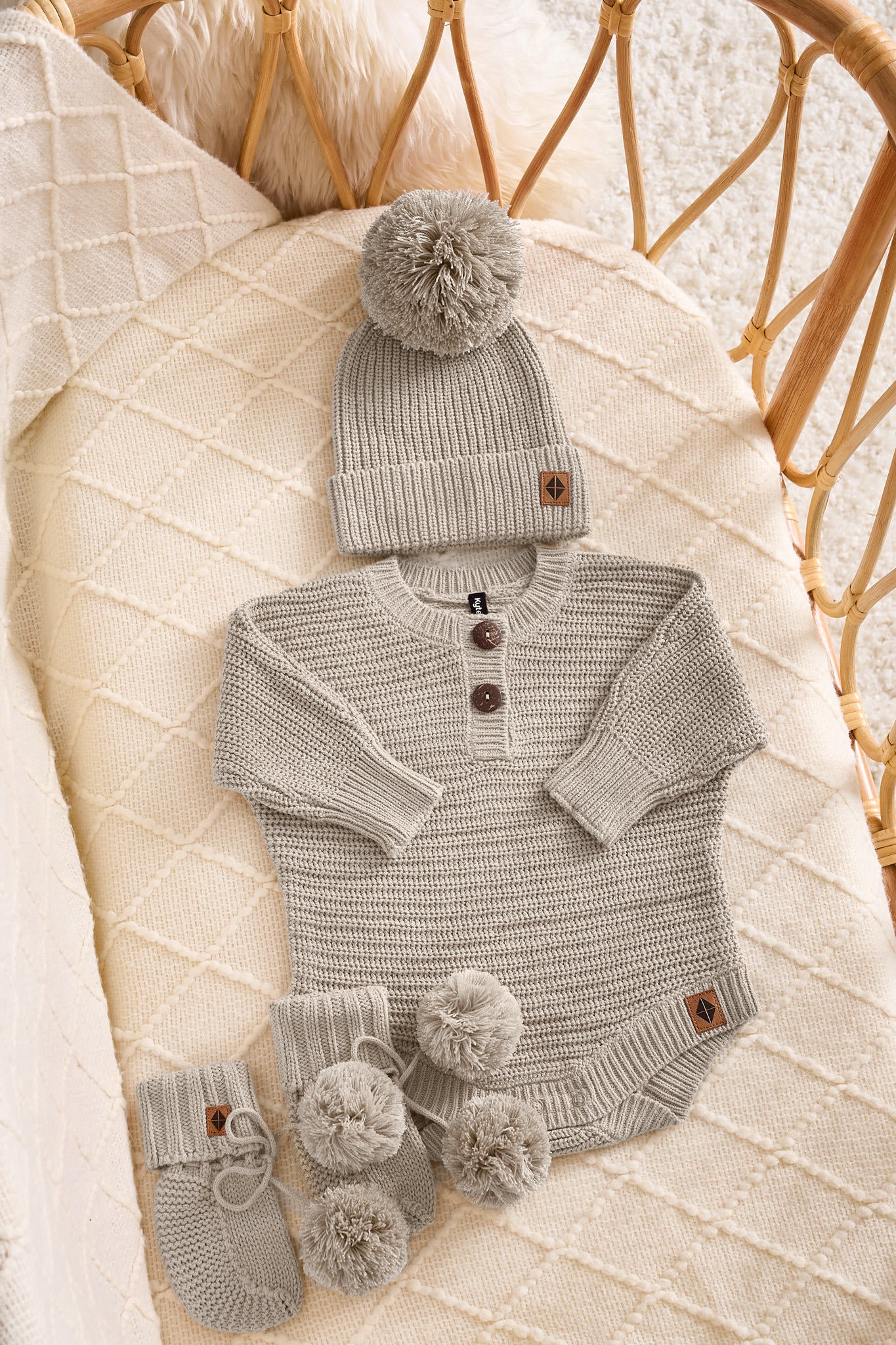 A baby knit outfit set in a wicker crib, featuring a light gray long-sleeve henley bodysuit with three buttons, a matching beanie with a pom-pom, and a pair of booties with pom-poms.