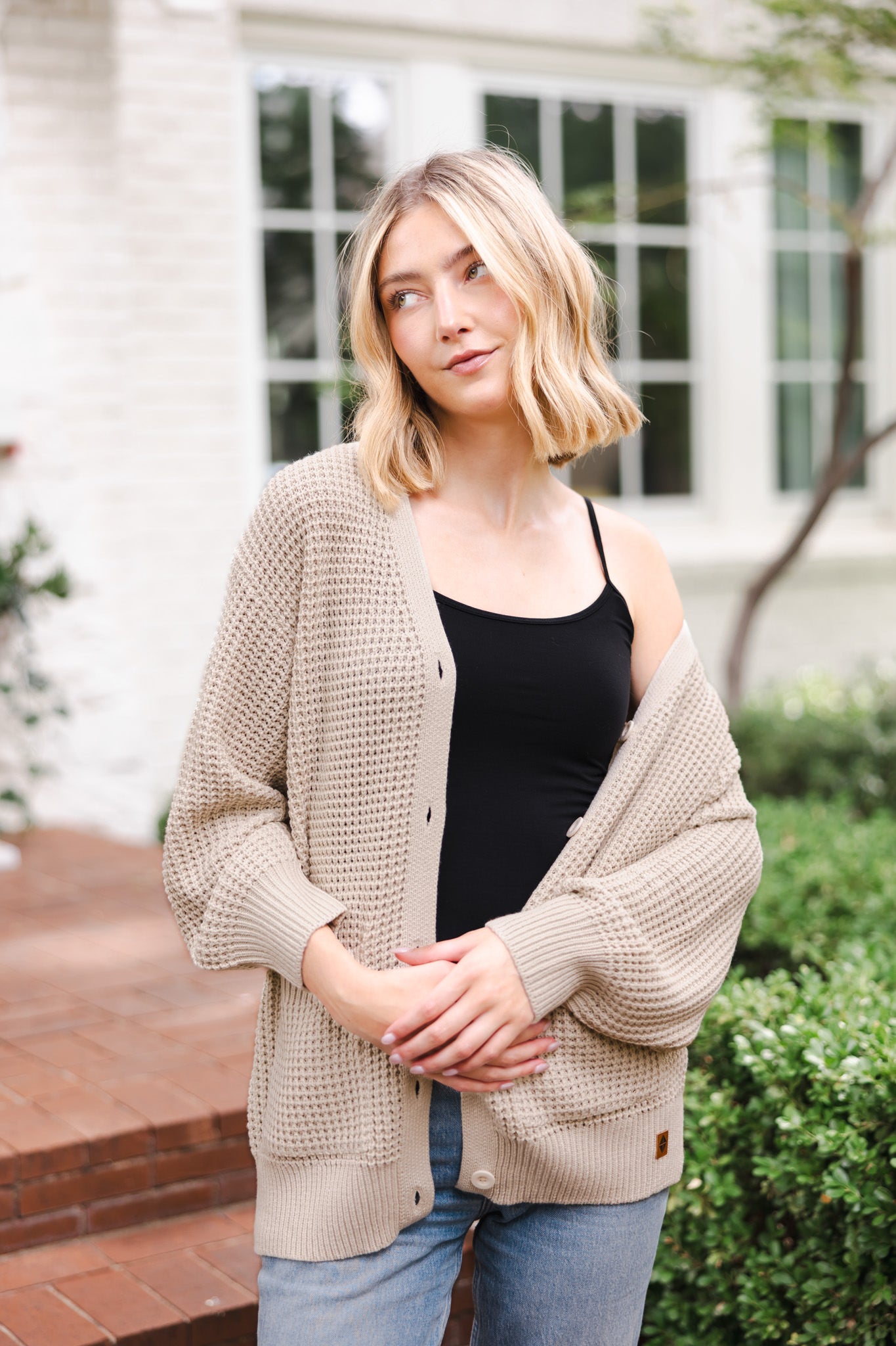 Modal posing outdoors wearing Chunky Knit Women's Waffle Cardigan in Almond over black camisole and blue jeans