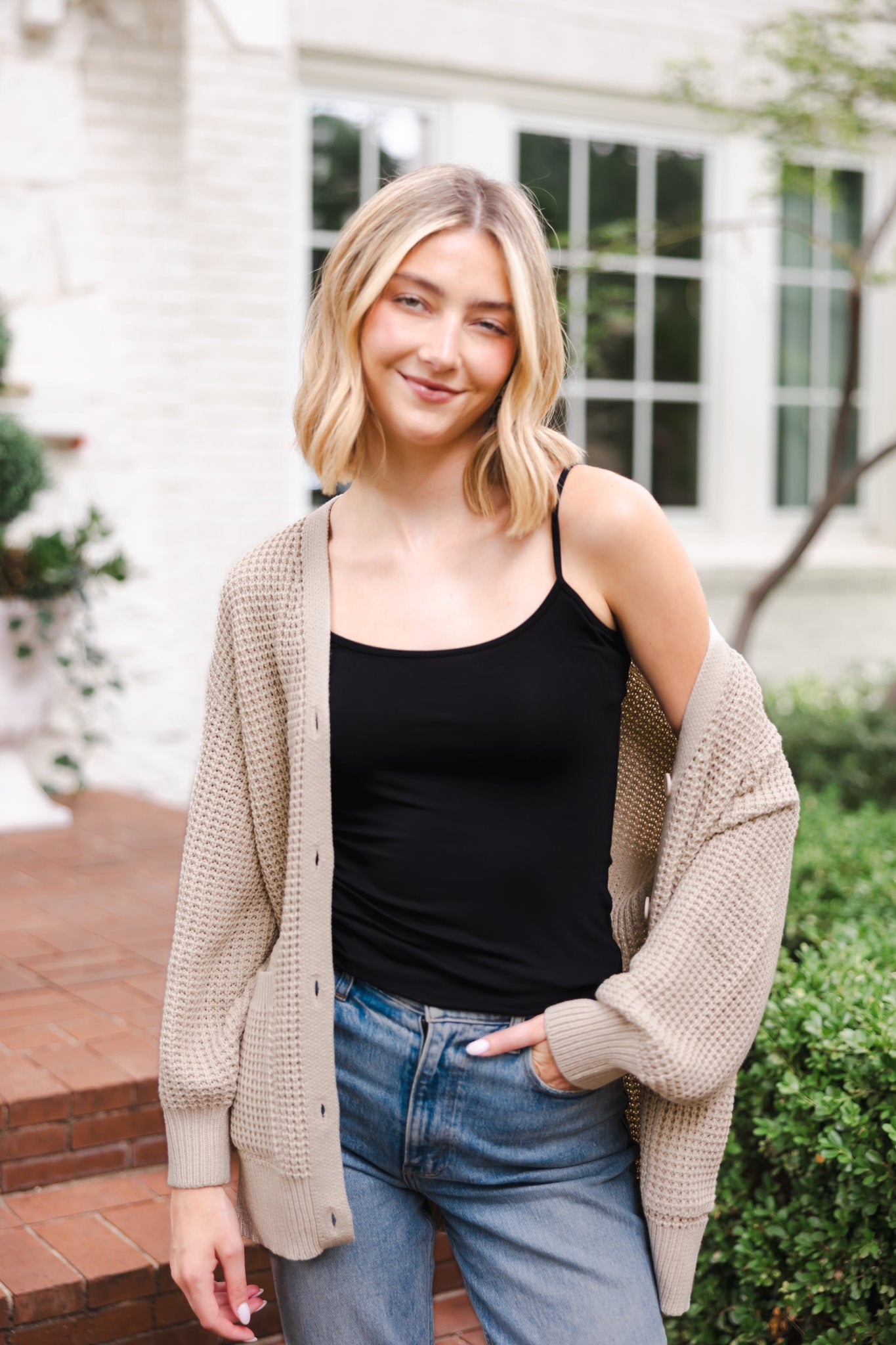 Modal smiling outdoors wearing Chunky Knit Women's Waffle Cardigan in Almond over black camisole and blue jeans