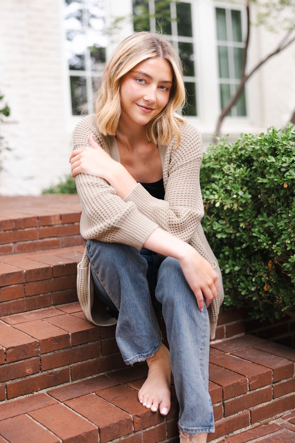 Modal sitting on stairs outdoors wearing Chunky Knit Women's Waffle Cardigan in Almond over black camisole and blue jeans